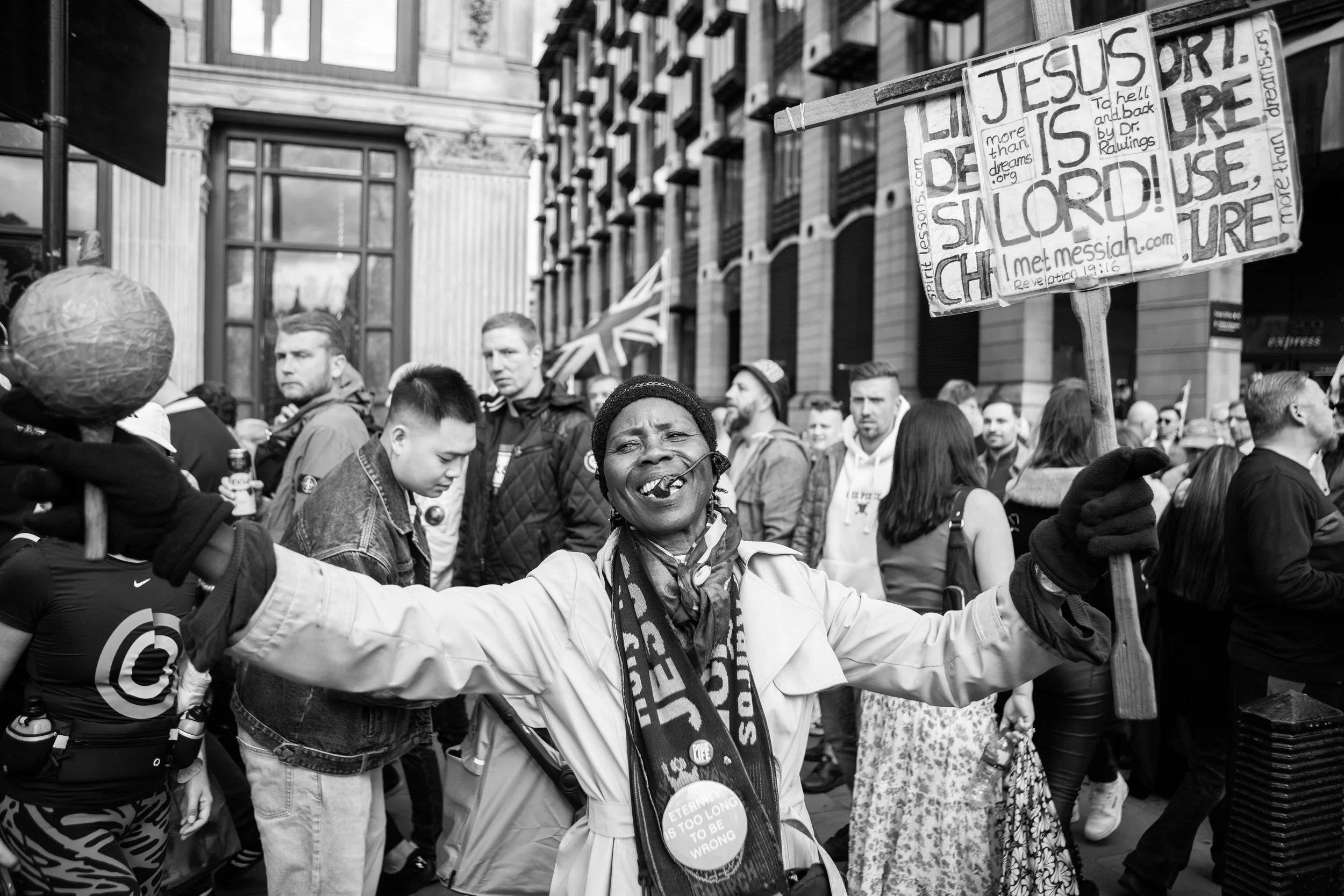 A woman joyfully participating in a protest, holding a sign with religious and political messages, surrounded by a diverse crowd on a city street.
