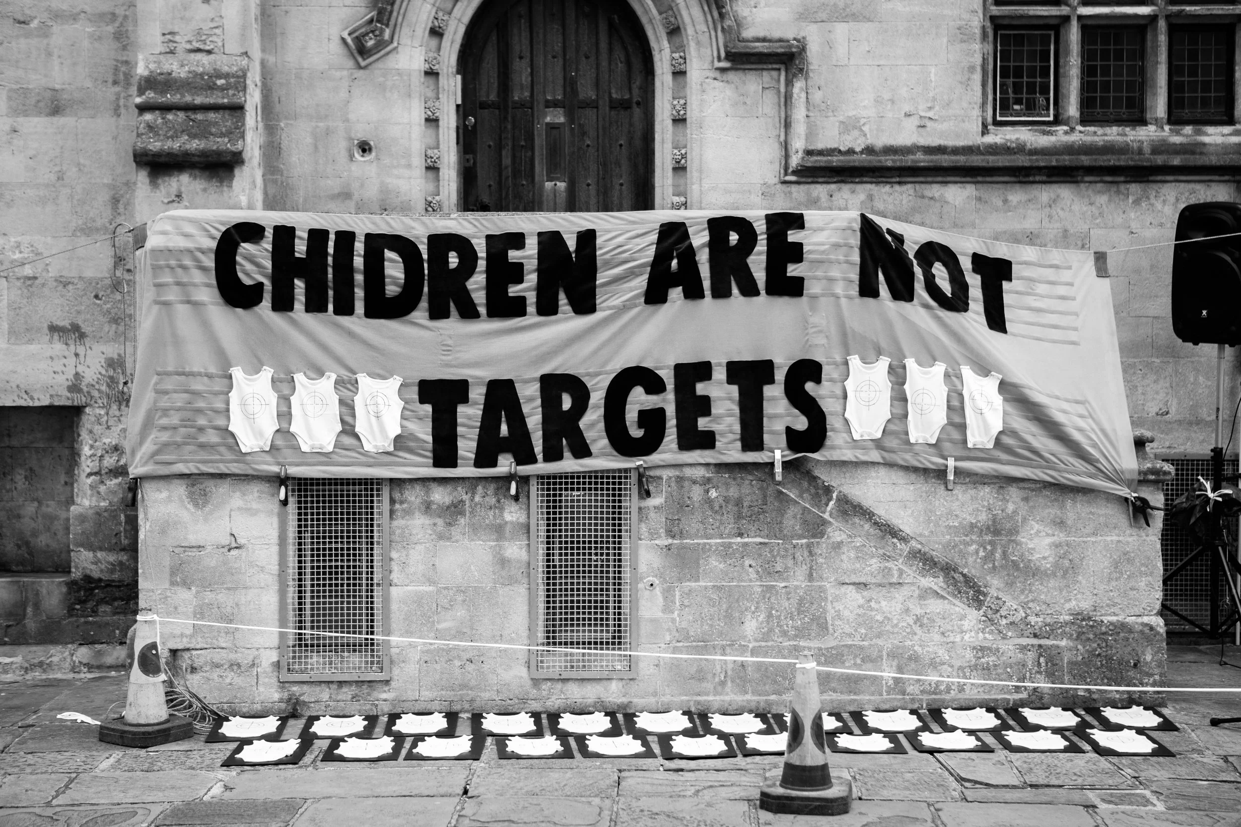 A protest display with a large banner that reads 'Children Are Not Targets,' accompanied by images of baby onesies with target symbols, set against an old building exterior.