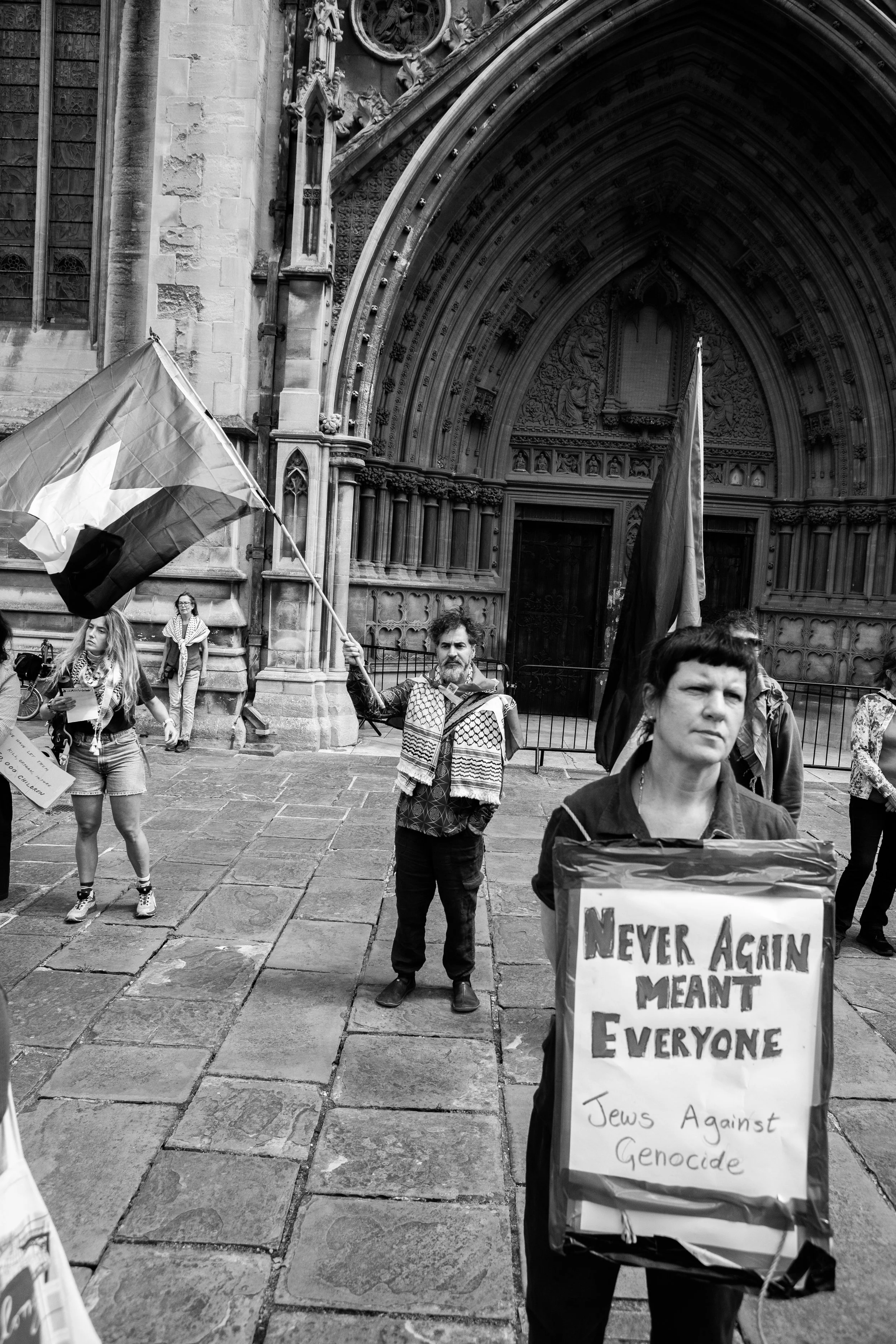 Protesters gathered outside a historic building, holding flags and signs. One woman in the foreground holds a sign that reads "Never Again Meant Everyone, Jews Against Genocide." A man in the background waves a flag.