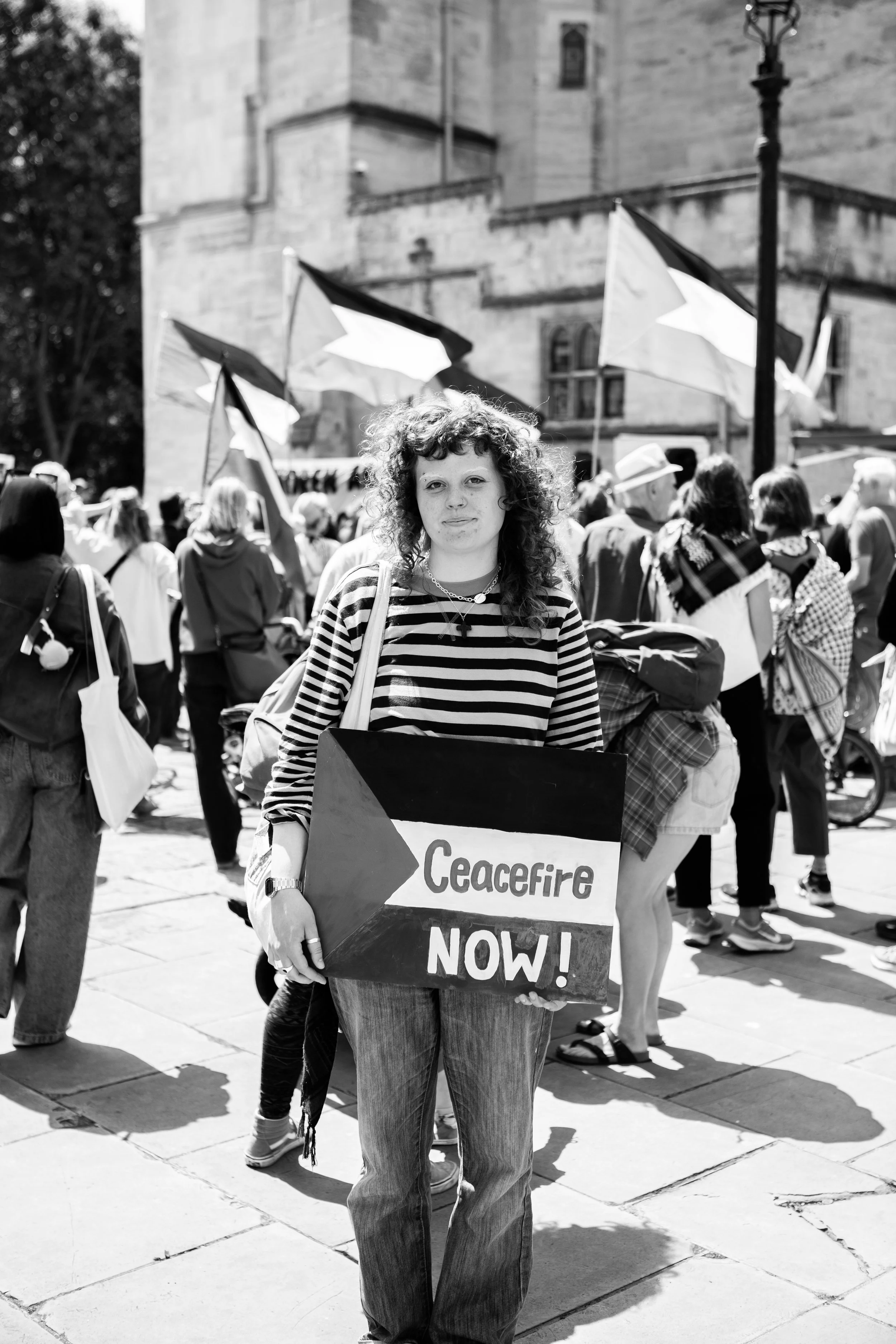 A woman with curly hair holding a sign that says 'Ceacefire NOW!' standing among a crowd during a protest.