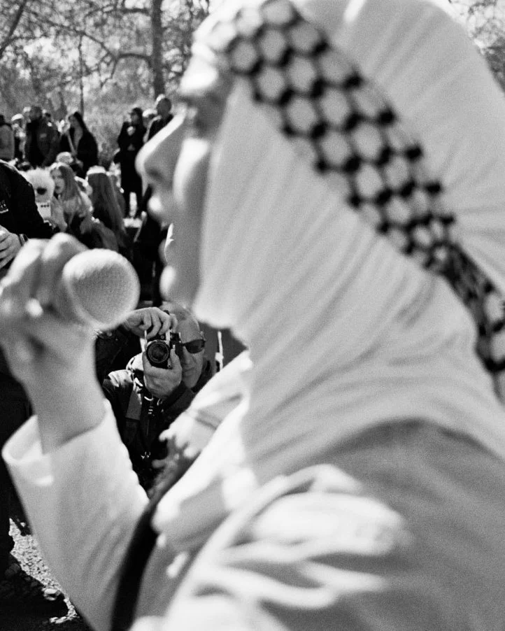 A person wearing a white headscarf with black and white patterns is speaking or singing into a microphone at an outdoor event, with a crowd and a photographer taking pictures in the background.