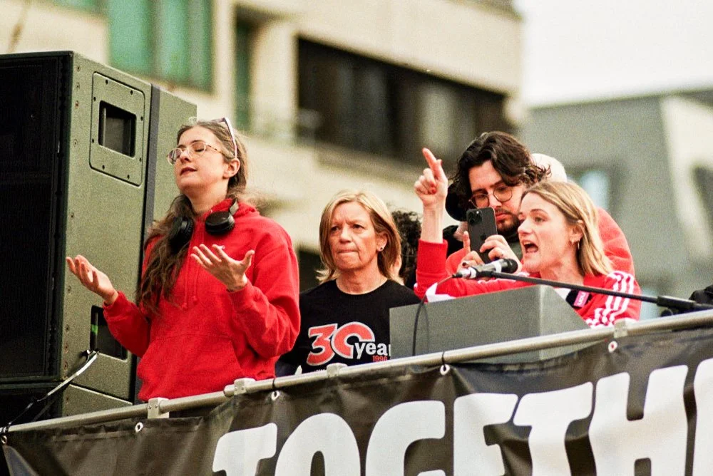 Four people standing behind a banner at a rally or protest, with two speaking and using a microphone, one recording with a phone, and one standing with eyes closed and hands raised.