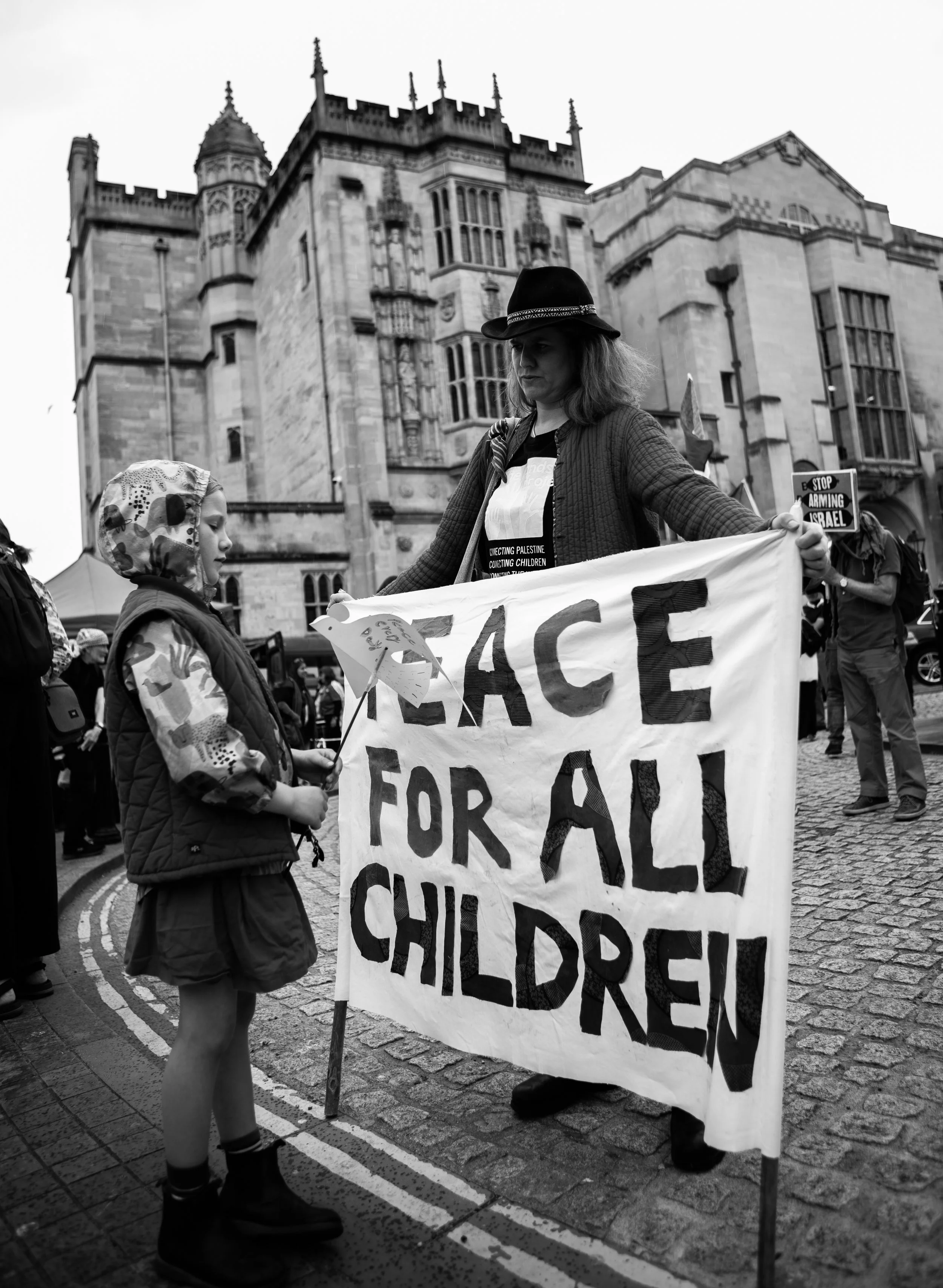 A protest scene with a woman and a girl holding a sign that reads 'PEACE FOR ALL CHILDREN' in front of a historic building. The woman is wearing a black hat and a jacket, and the girl has a hooded jacket and is holding a small dove-shaped peace symbo