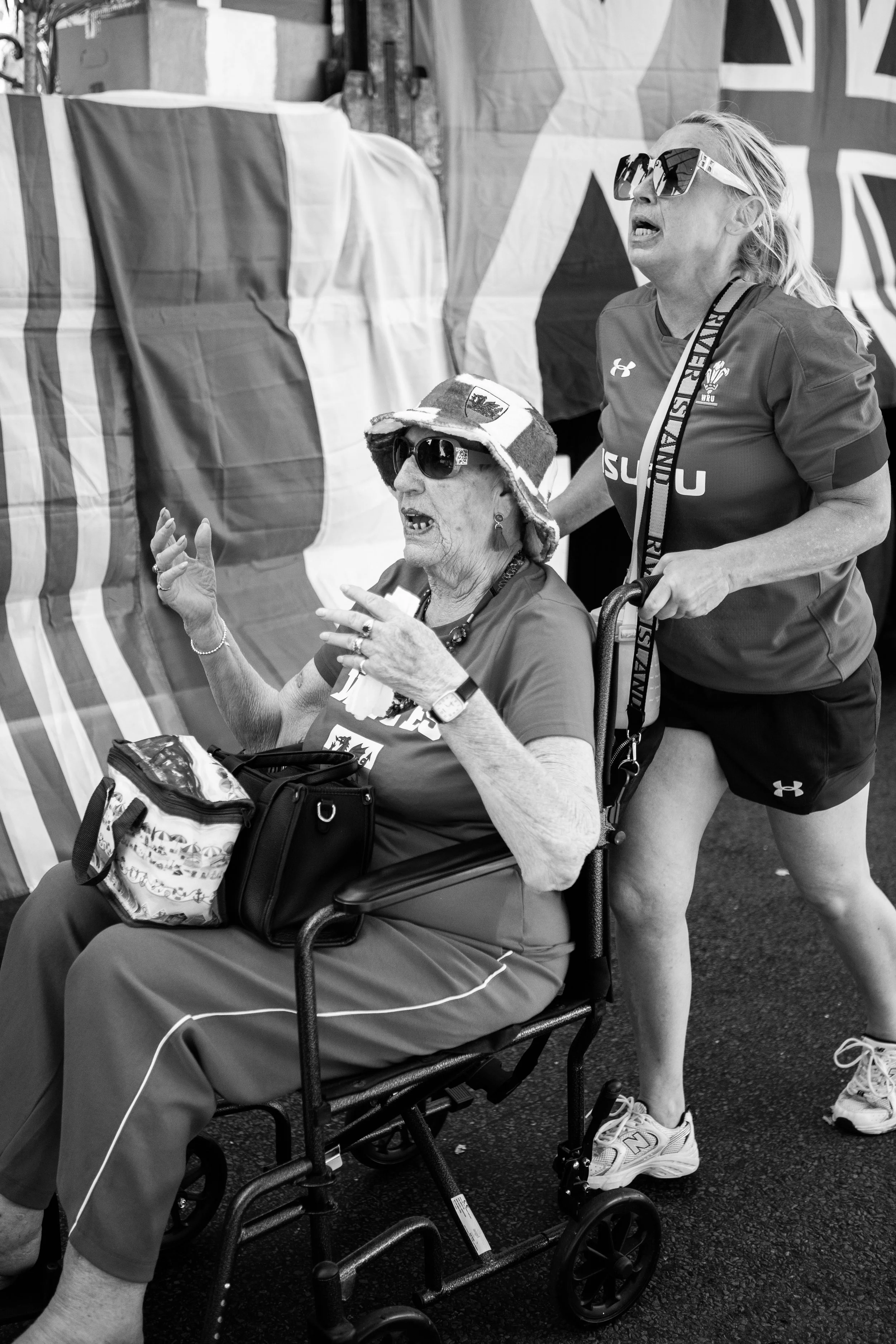 An elderly woman in a wheelchair wearing sunglasses, a hat with patriotic symbols, and a sports shirt, being pushed by a younger woman in sportswear. They are at a patriotic or sports event, with flags in the background.
