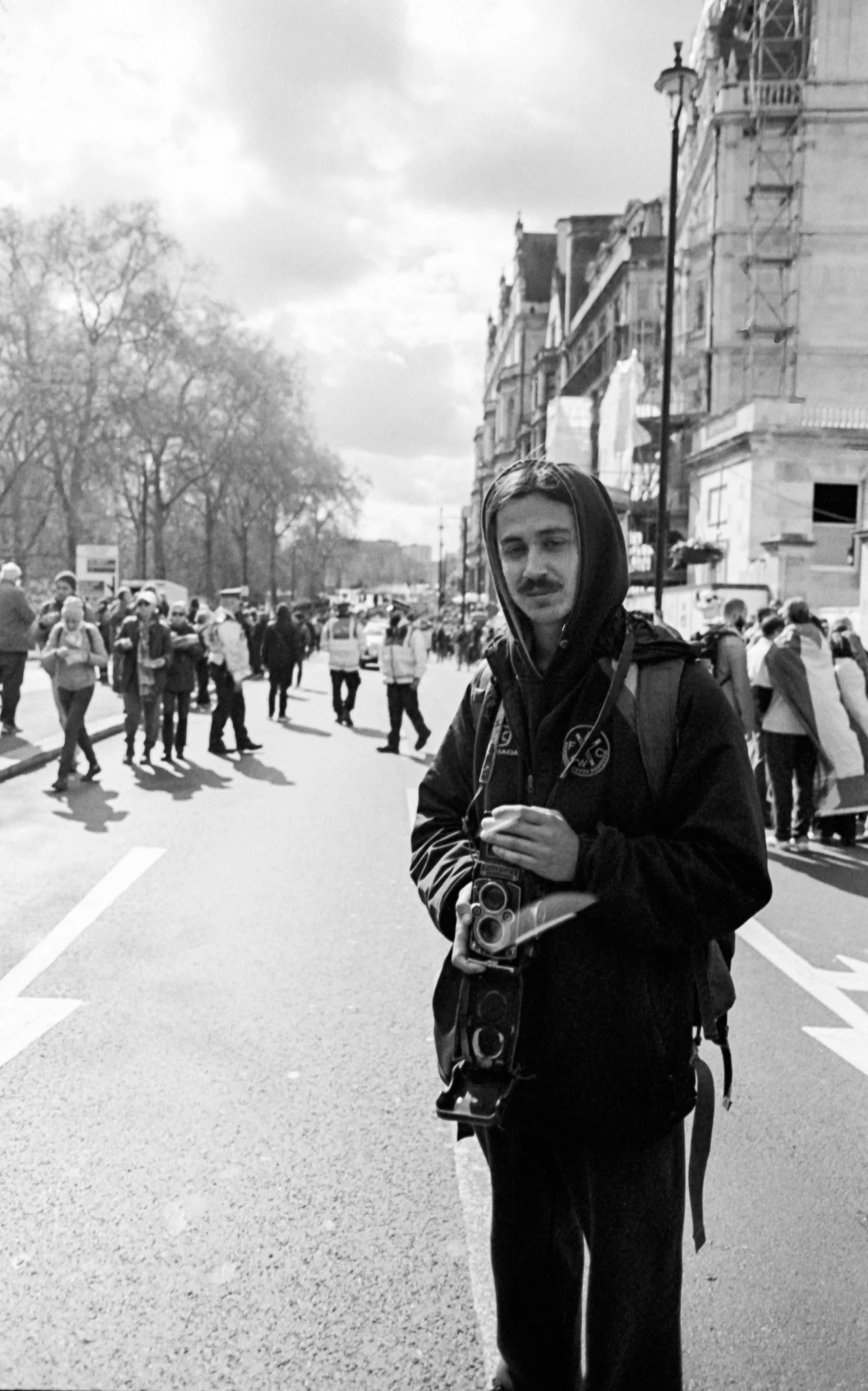 A young man holding a camera, wearing a hoodie and jacket, standing on a city street with a crowd of people in the background.
