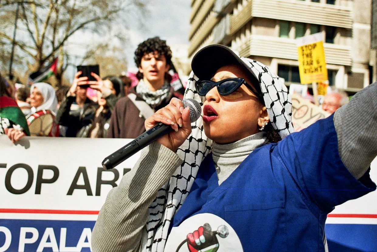Woman wearing sunglasses and a keffiyeh scarf speaking into a microphone at a protest or rally, with a crowd and protest signs behind her.