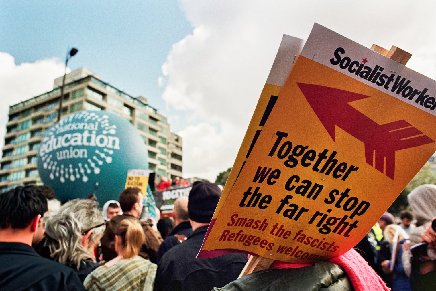 Protest crowd holding signs, prominently one sign reads 'Together we can stop the far right. Smash the fascists. Refugees welcome.' in front of a large blue balloon with the words 'National Education Union' during a demonstration outside a building.