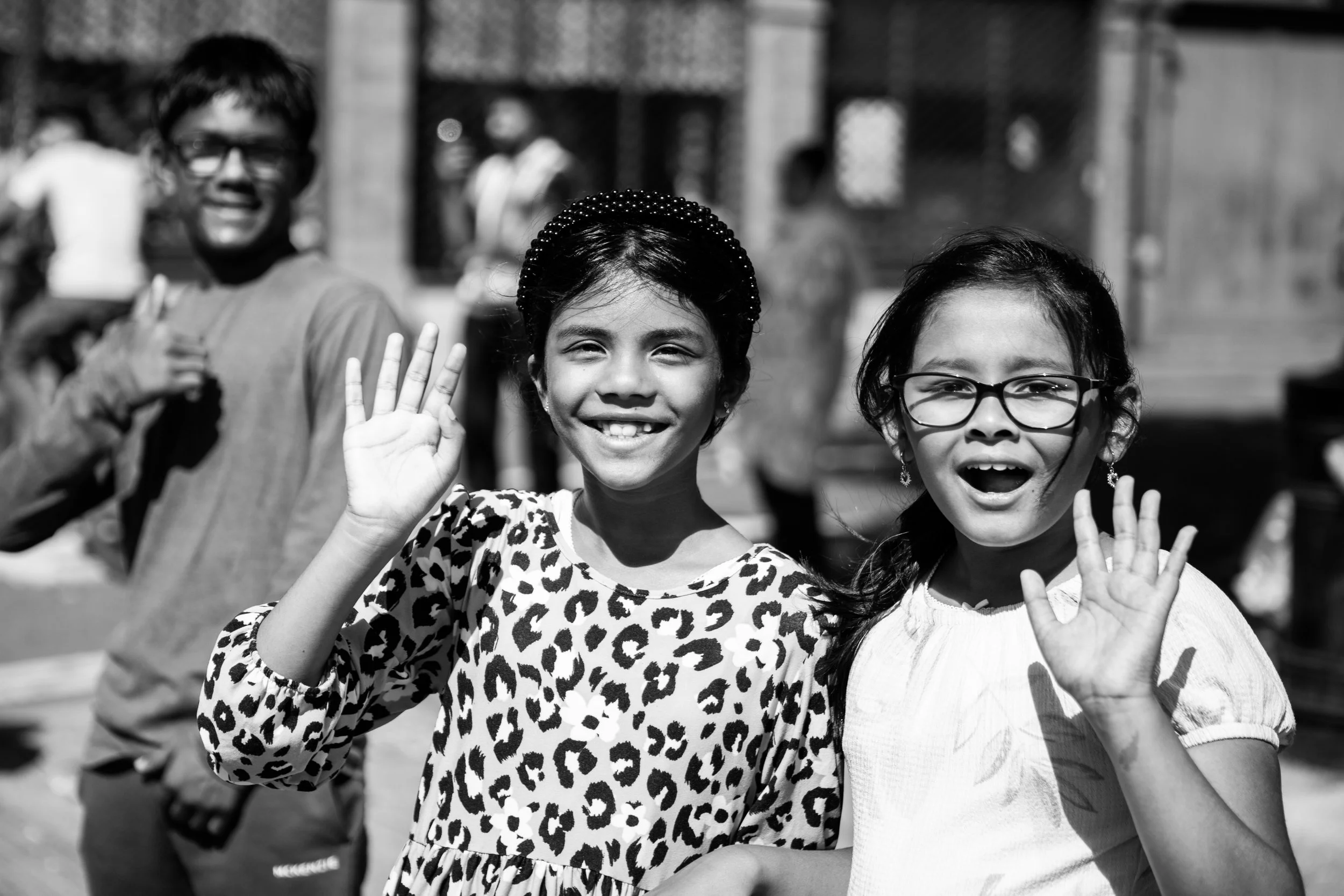 Two young girls smiling and waving at the camera, standing outdoors, with an older man in the background.