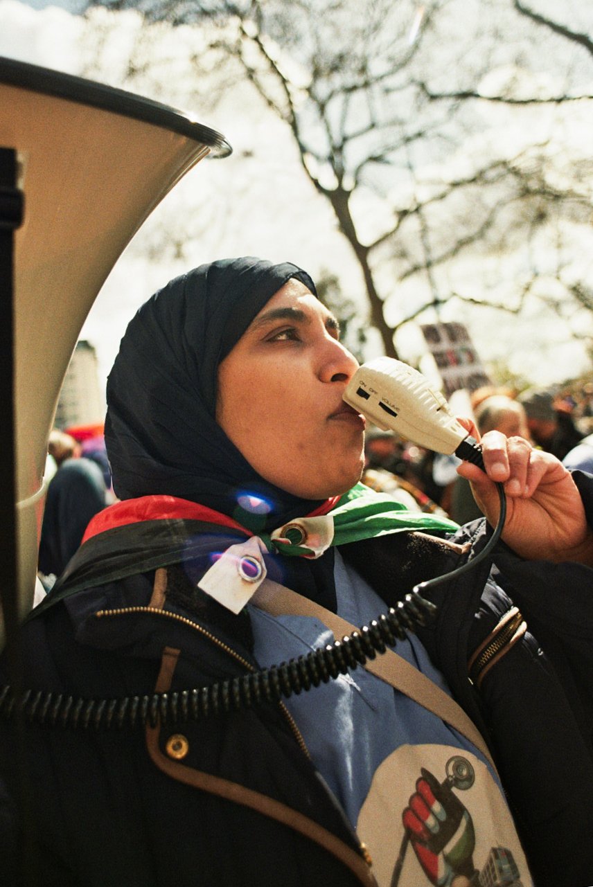 A woman wearing a black hijab and a protest or rally badge, speaking into a handheld microphone, outdoors with trees and a crowd in the background.