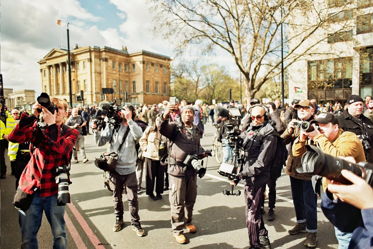 A group of photographers and journalists with cameras in a crowded outdoor setting with a large building and a tree in the background.
