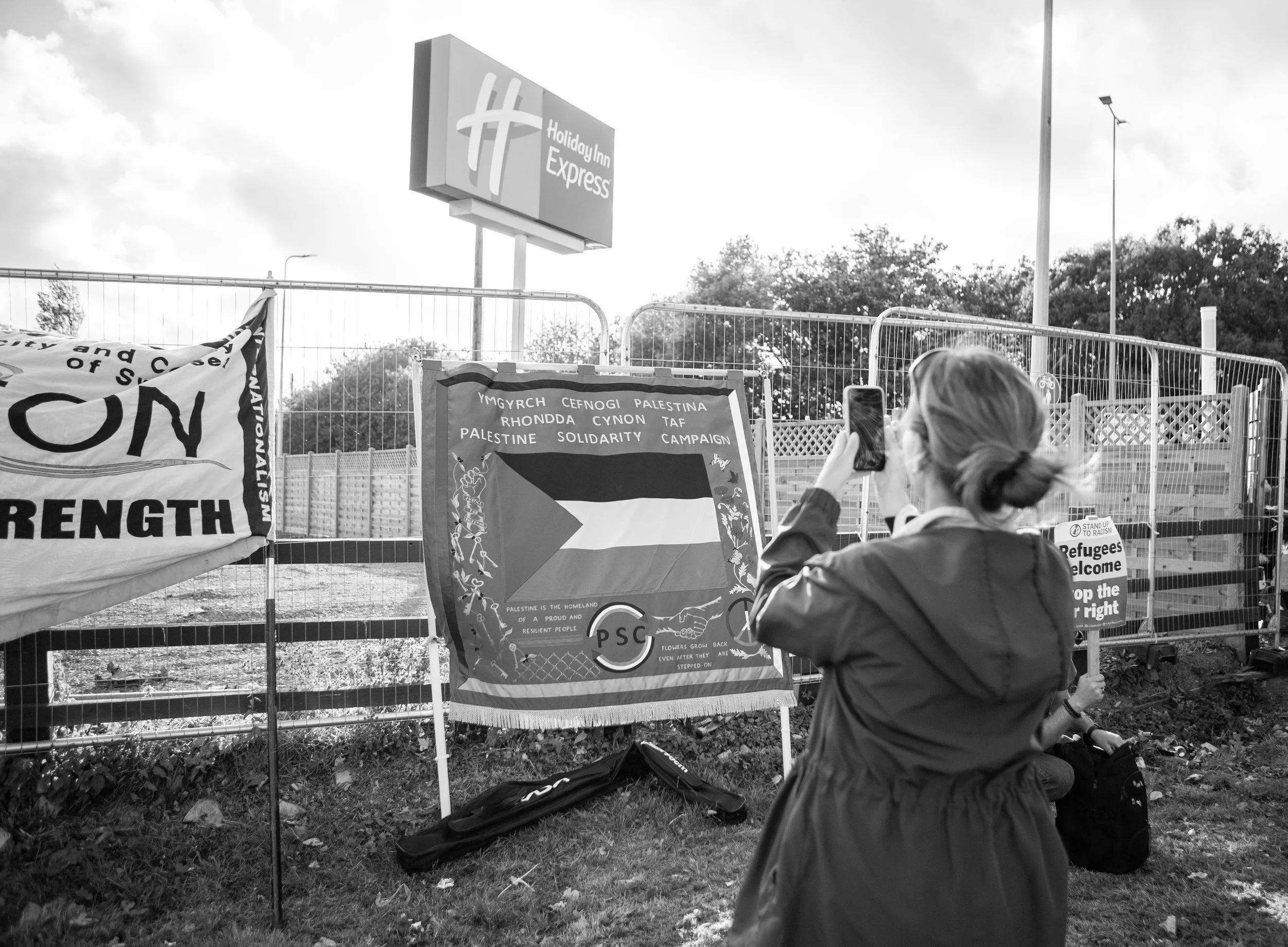 A woman taking a photo with her phone of protest banners at a fence near a Holiday Inn Express, Cardiff.