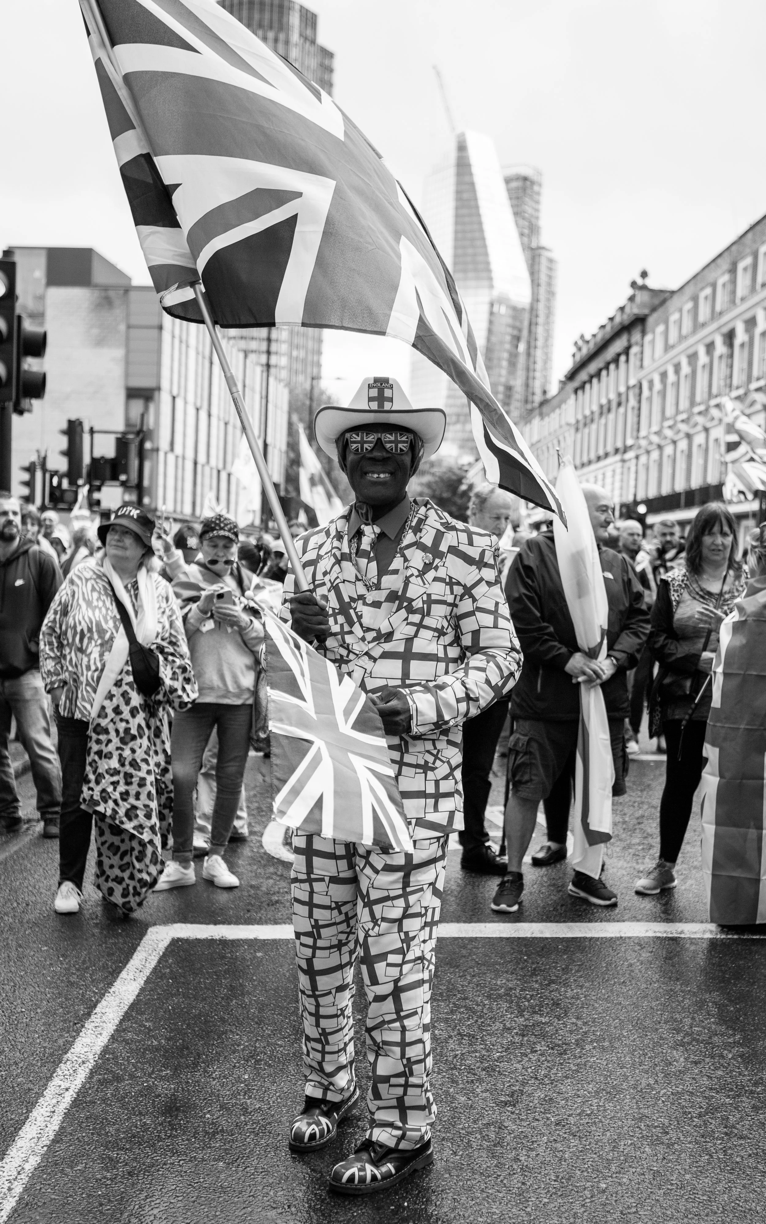 A man dressed in British-themed clothing holding a Union Jack flag at a street protest or parade, with a crowd and city buildings in the background.