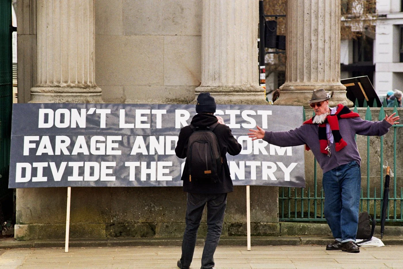 A man with a gray beard and hat gestures with his arms extended while speaking to another person in front of a sign that reads, "DON'T LET R Sponsor PASTE AND FORM DIVIDE THE COUNTRY." The scene is outdoors near stone columns and a fence.