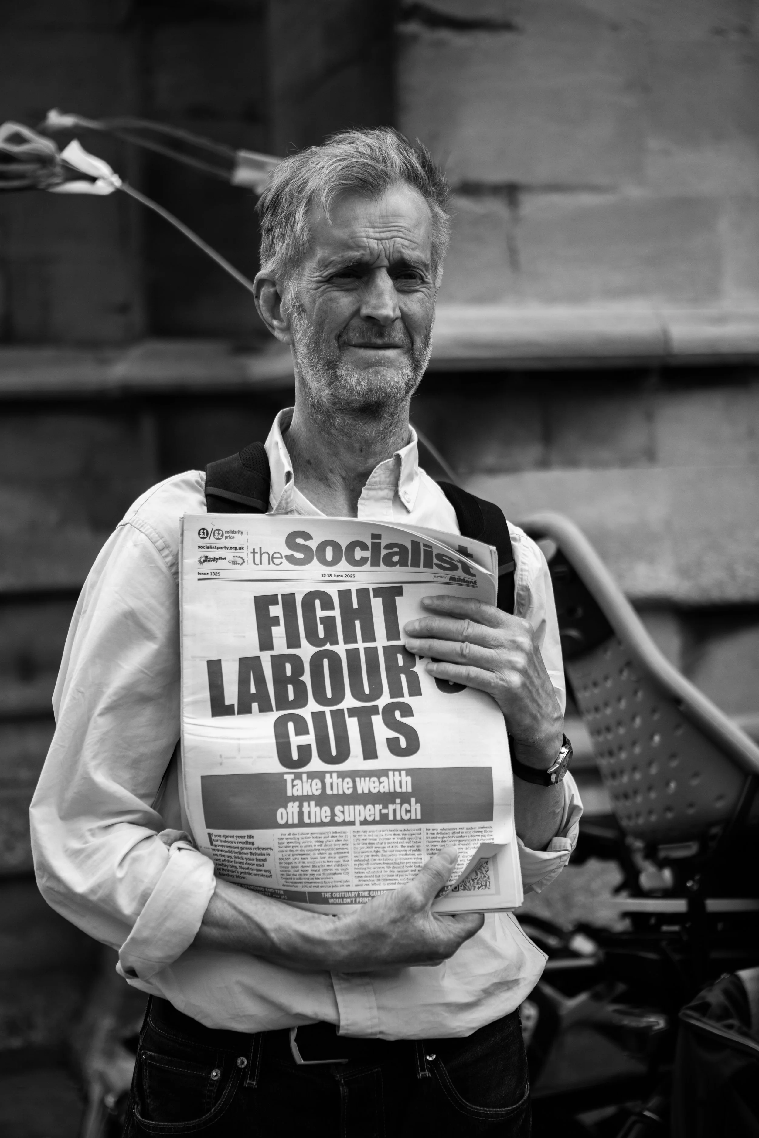 A man with gray hair and beard holding a newspaper titled 'The Socialist' with a headline about fighting labor cuts and taxing the wealthy, standing outdoors.
