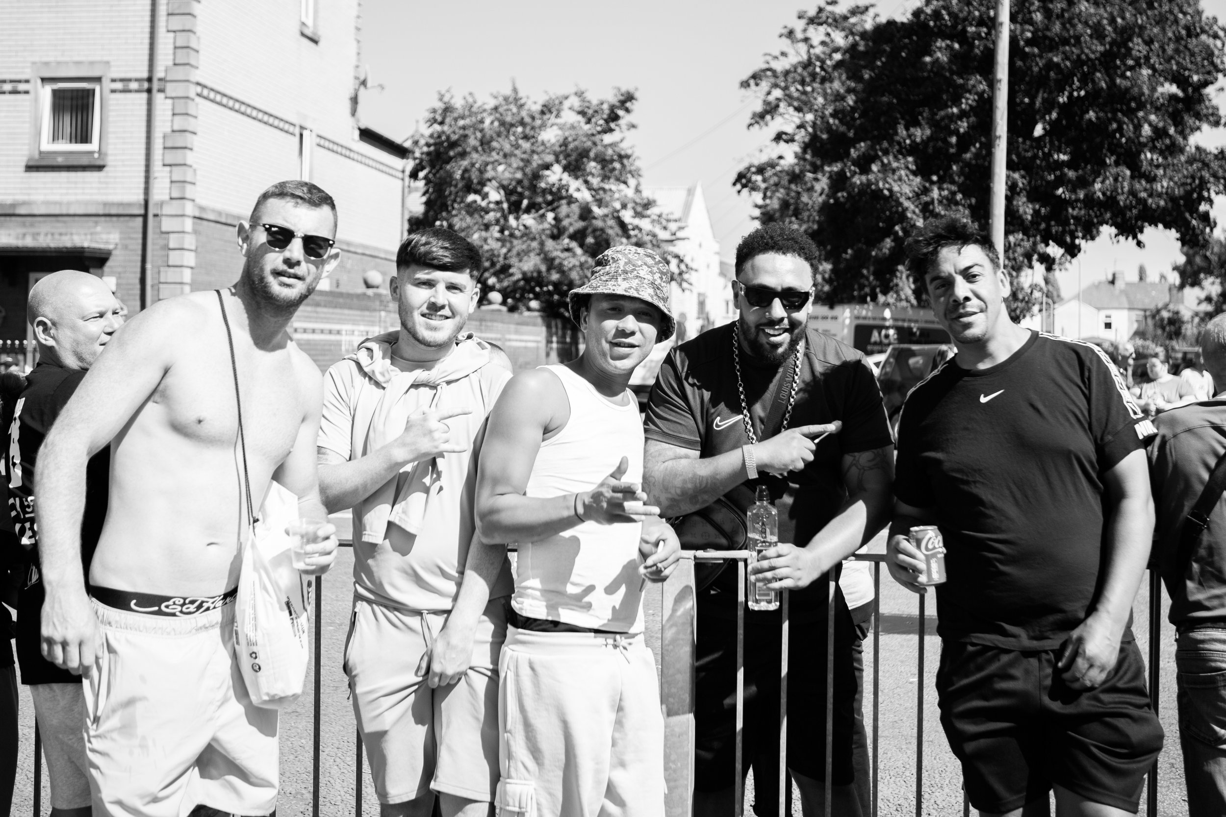 Five men posing outdoors near a metal barrier on a sunny day, some holding drinks, with trees and buildings in the background.