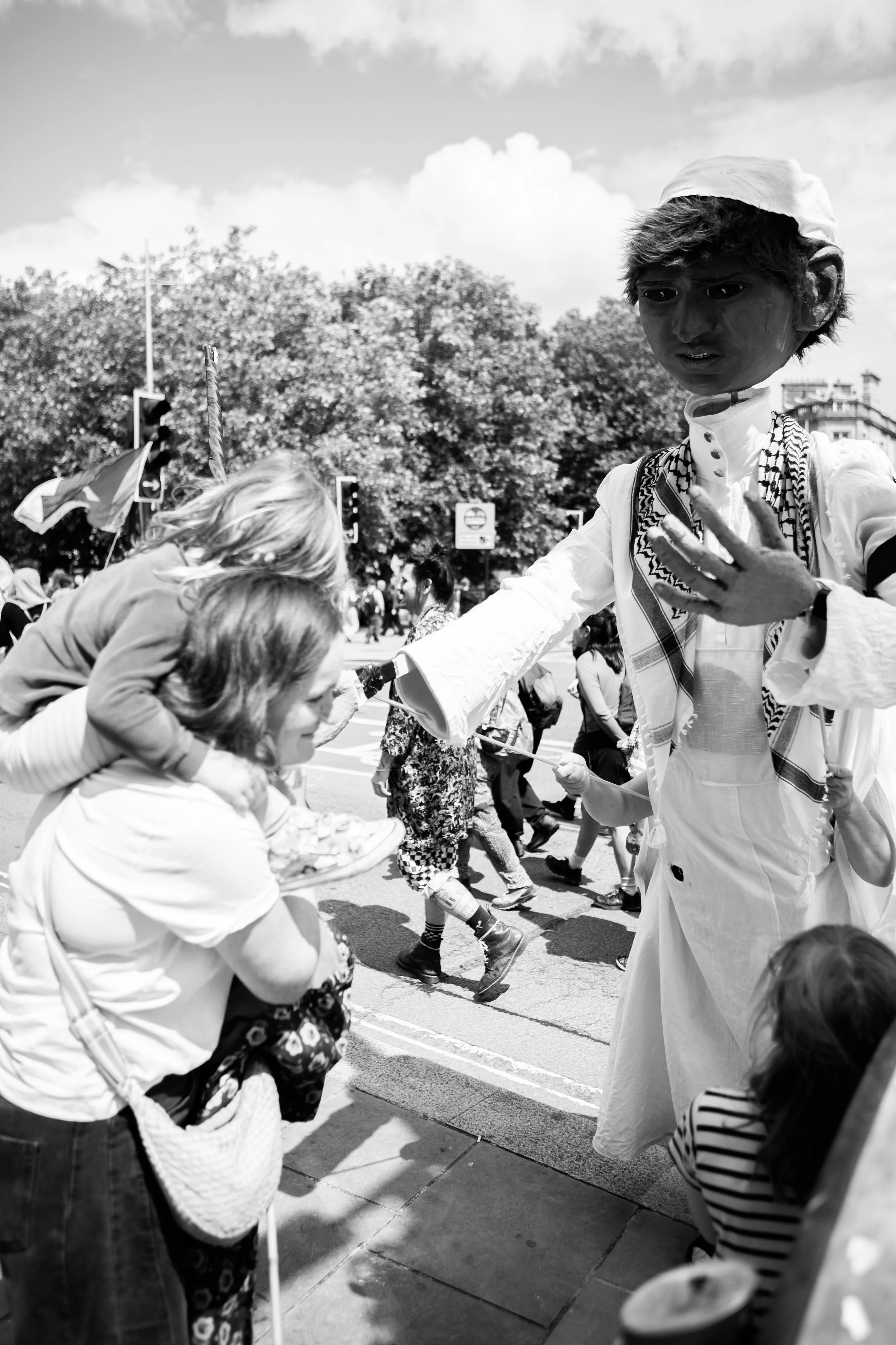 Street scene with children and an adult interacting with a street performer dressed in traditional clothing during a parade or festival, with trees and traffic lights in the background.