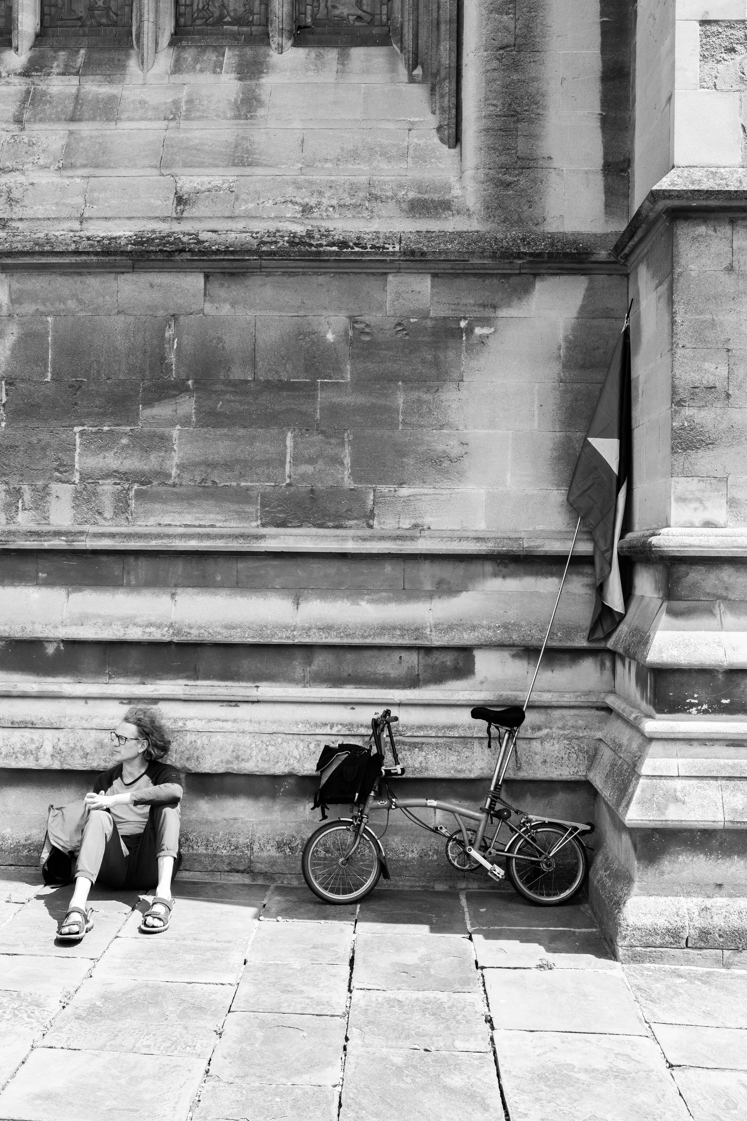 A woman sitting on the ground beside a bicycle with a flag attached, against a stone wall of a building.