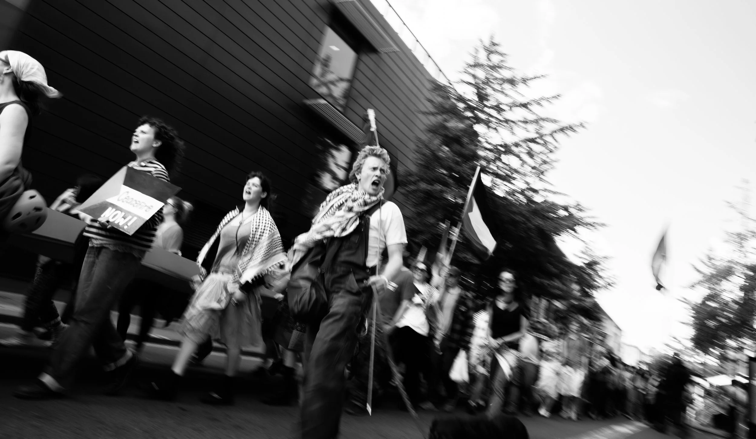 Black and white photo of a group of protesters marching. One woman with curly hair and an anti-war scarf is shouting passionately, holding a cane. Several flags are visible, and there are people walking along the street in front of a modern building 