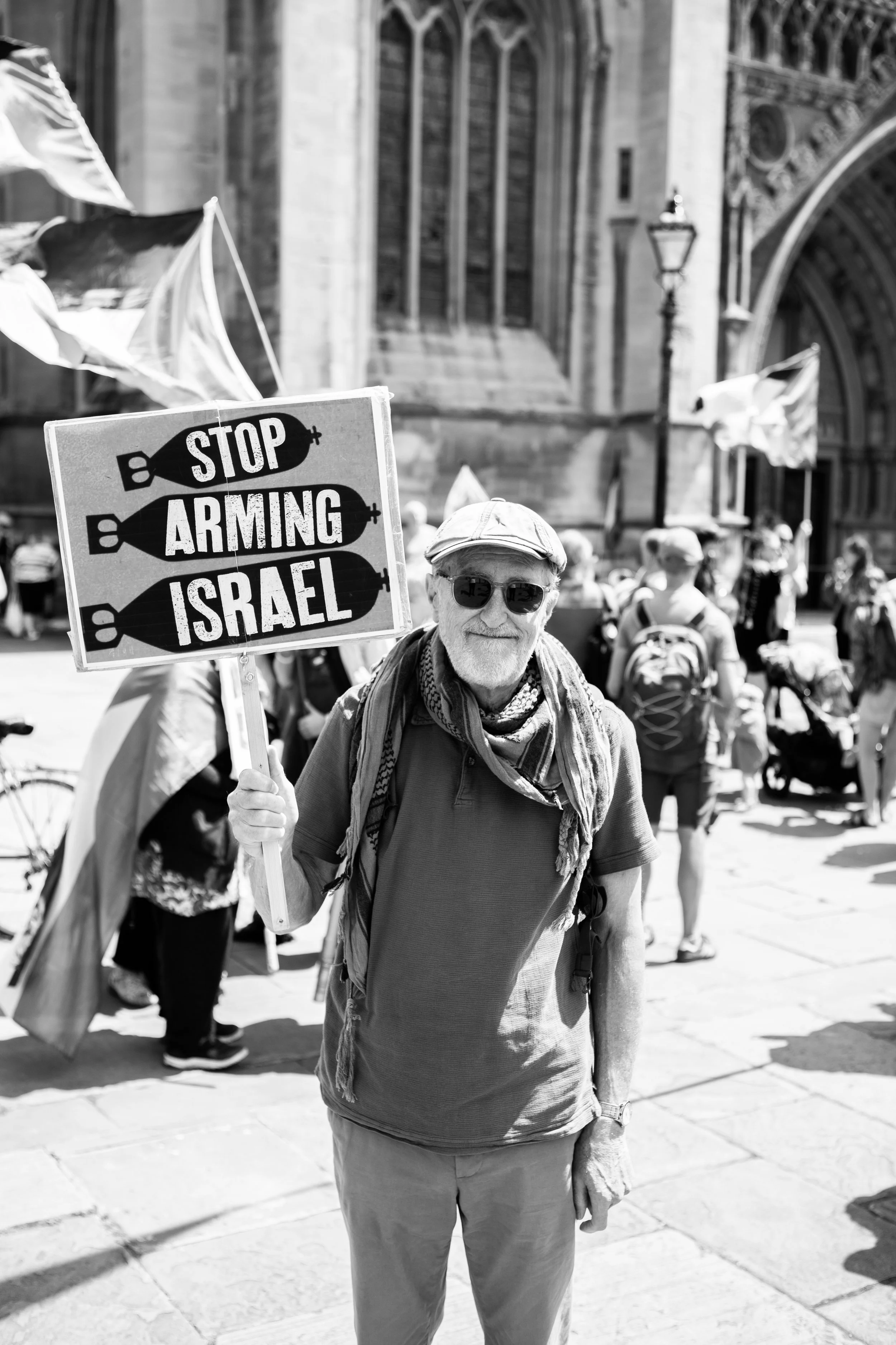 Older man wearing sunglasses, a cap, and a scarf holds a sign protesting against arms used in Israel, with a background of protesters and a historic building.