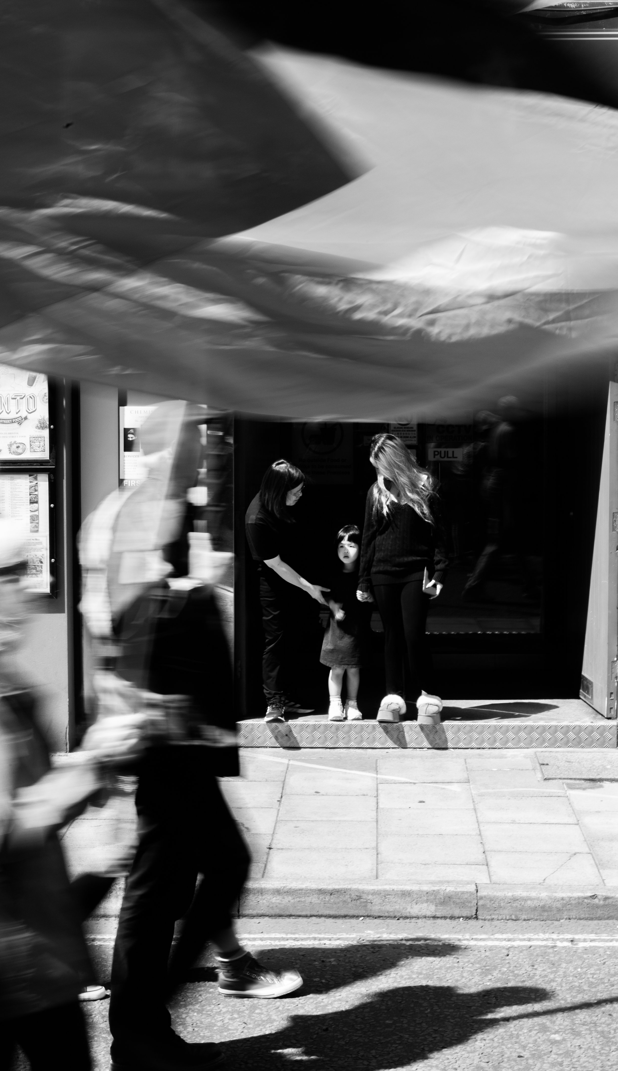 A black and white photograph showing a woman with long hair, a woman with glasses, and a young girl standing at the entrance of a building, likely asking for help, while blurred pedestrians walk past on the sidewalk.