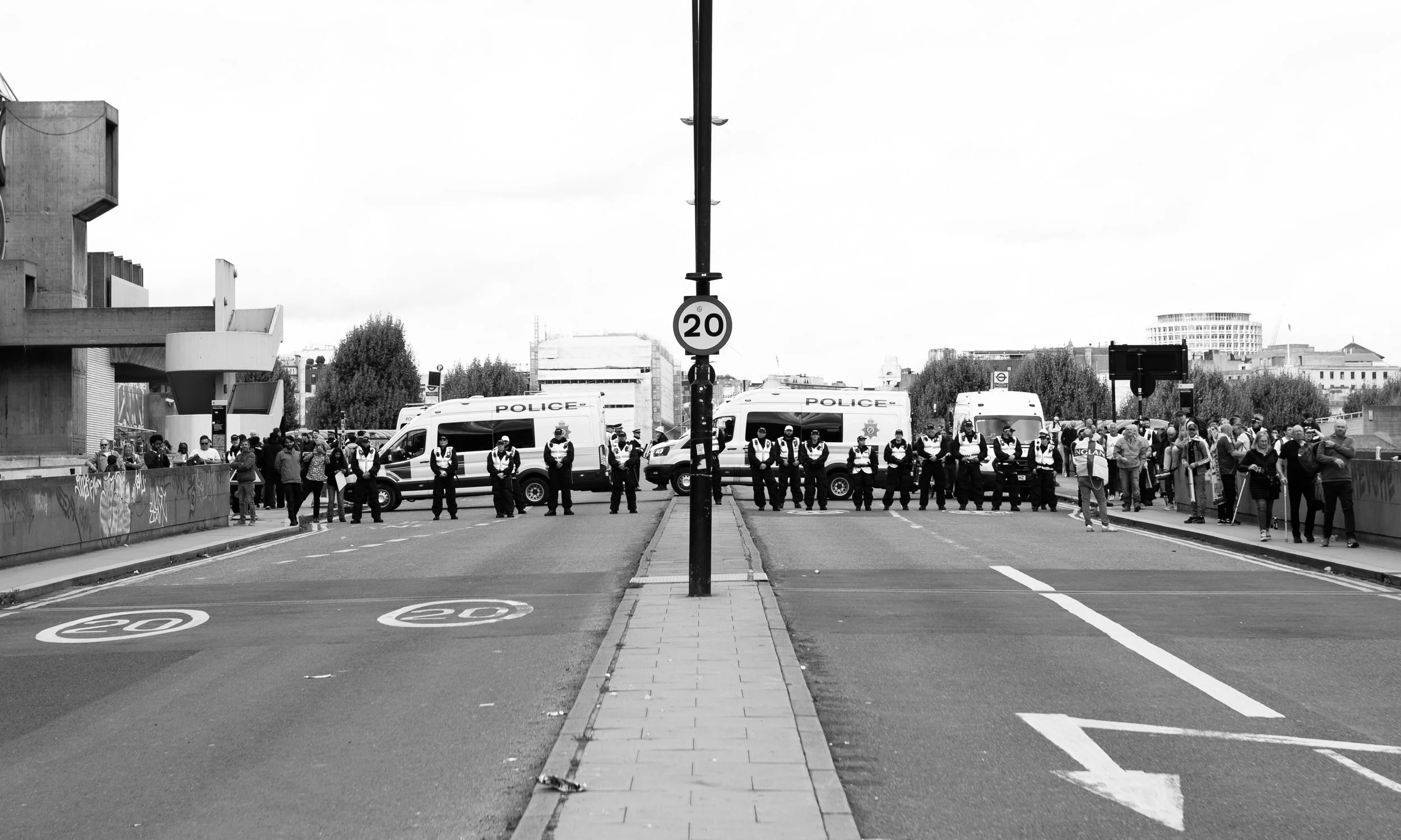 Black and white photo of police vans and officers forming a barrier on the street, with people gathered behind them on the sidewalk, in an urban environment.