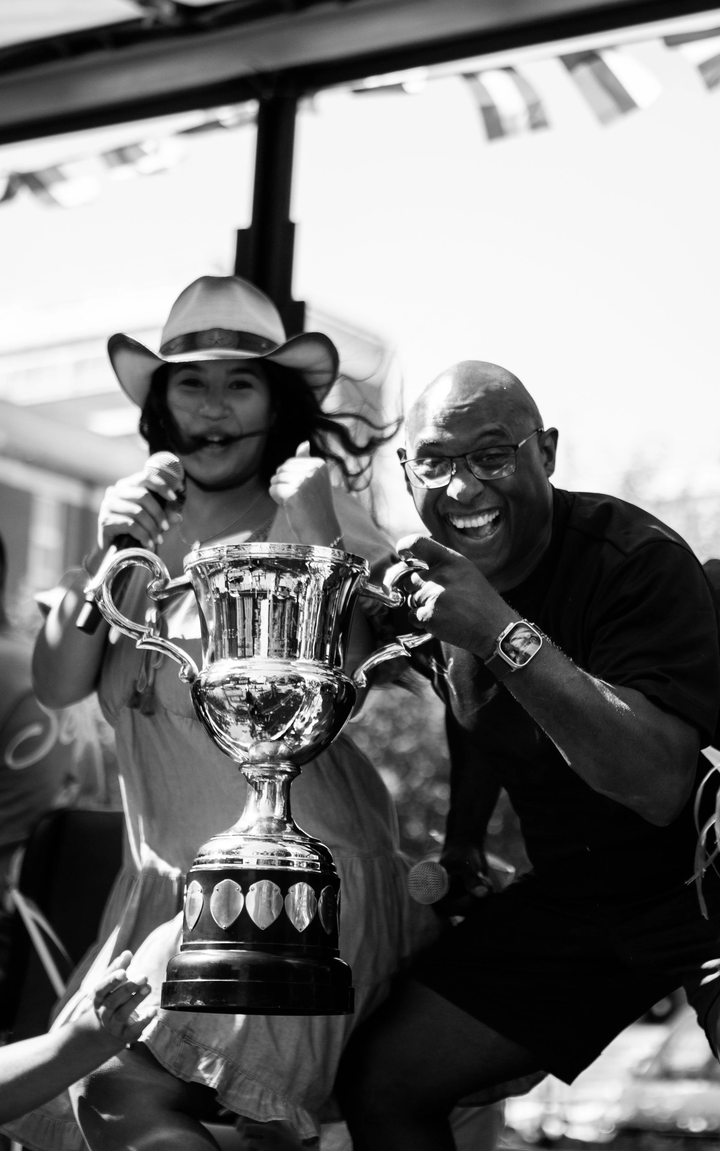 Two people celebrating outdoors with a large trophy, smiling and pointing, one wearing a cowboy hat and holding a microphone, the other pointing and smiling broadly.
