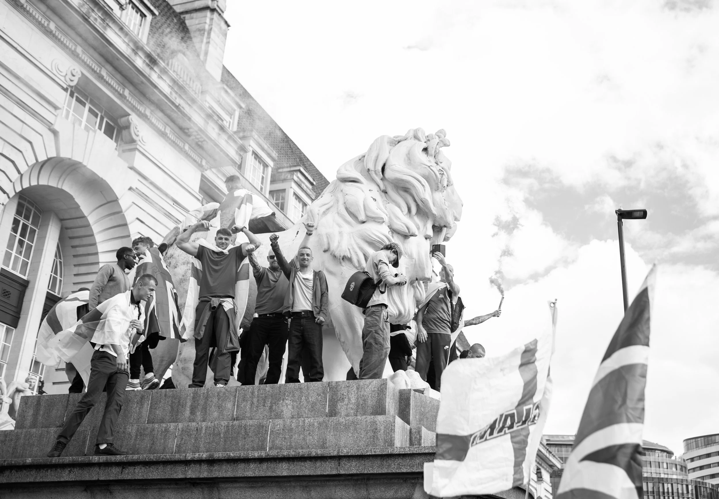 A group of people standing on a monument featuring a large lion's head sculpture, some holding flags, with a man smoking a cigarette, and others taking photos, in an urban setting with buildings in the background.