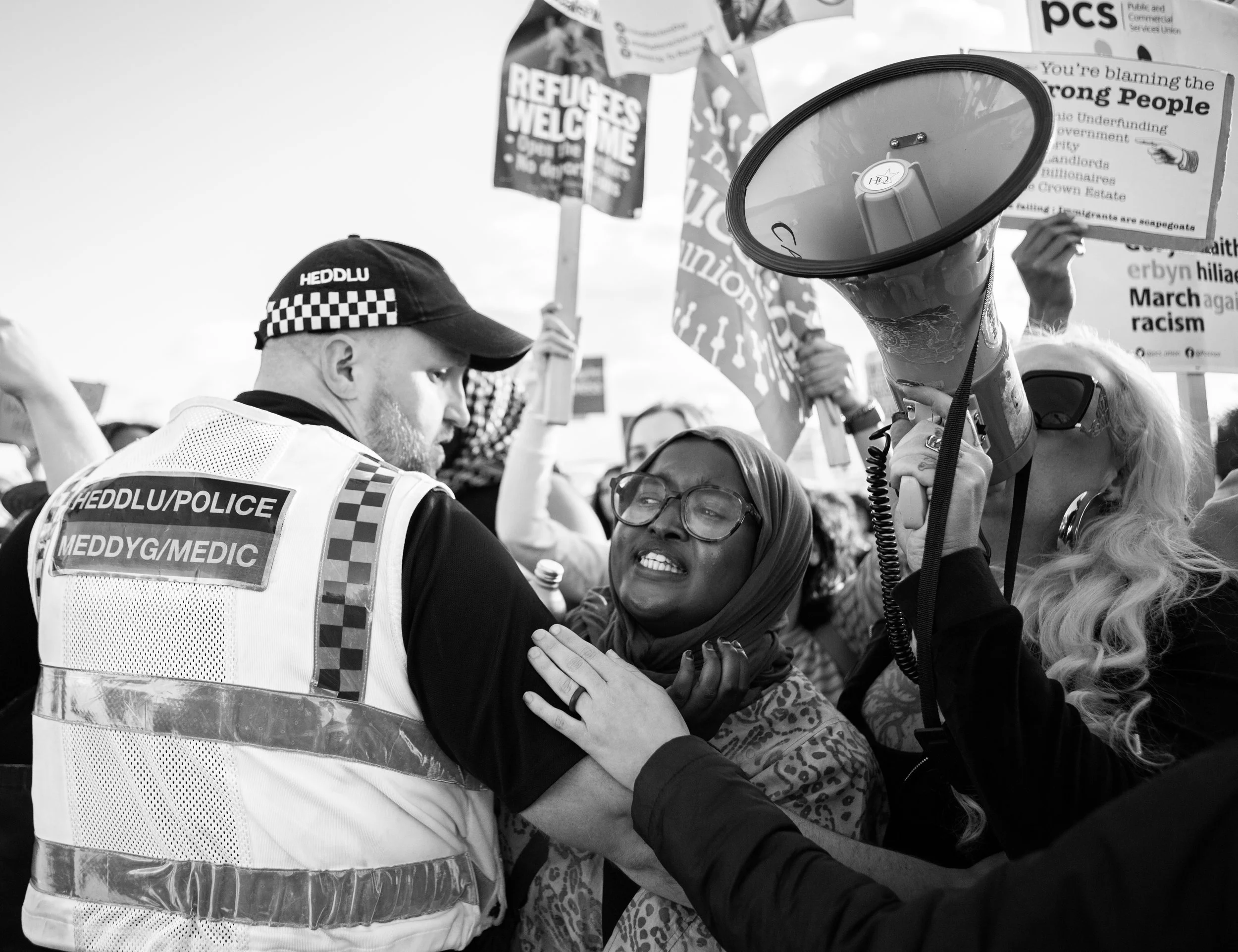 A woman wearing glasses and a headscarf is arguing with a police officer in a high-visibility vest. The woman is holding a megaphone, and there are protest signs held by other people in the background in a crowd of demonstrators.