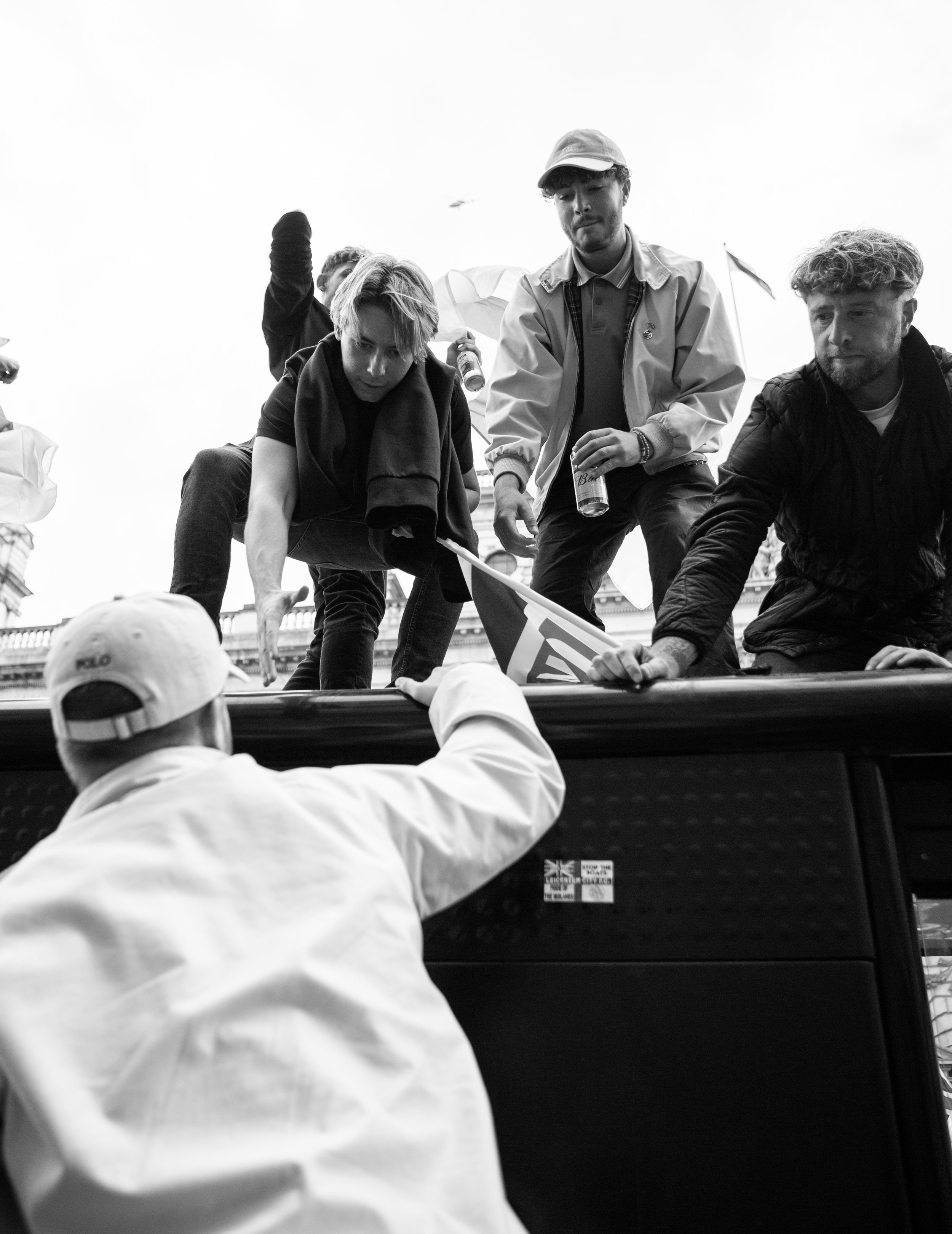 People participating in a protest or rally, some reaching into a vehicle with others on top of it, holding flags and beverages, in an urban setting.
