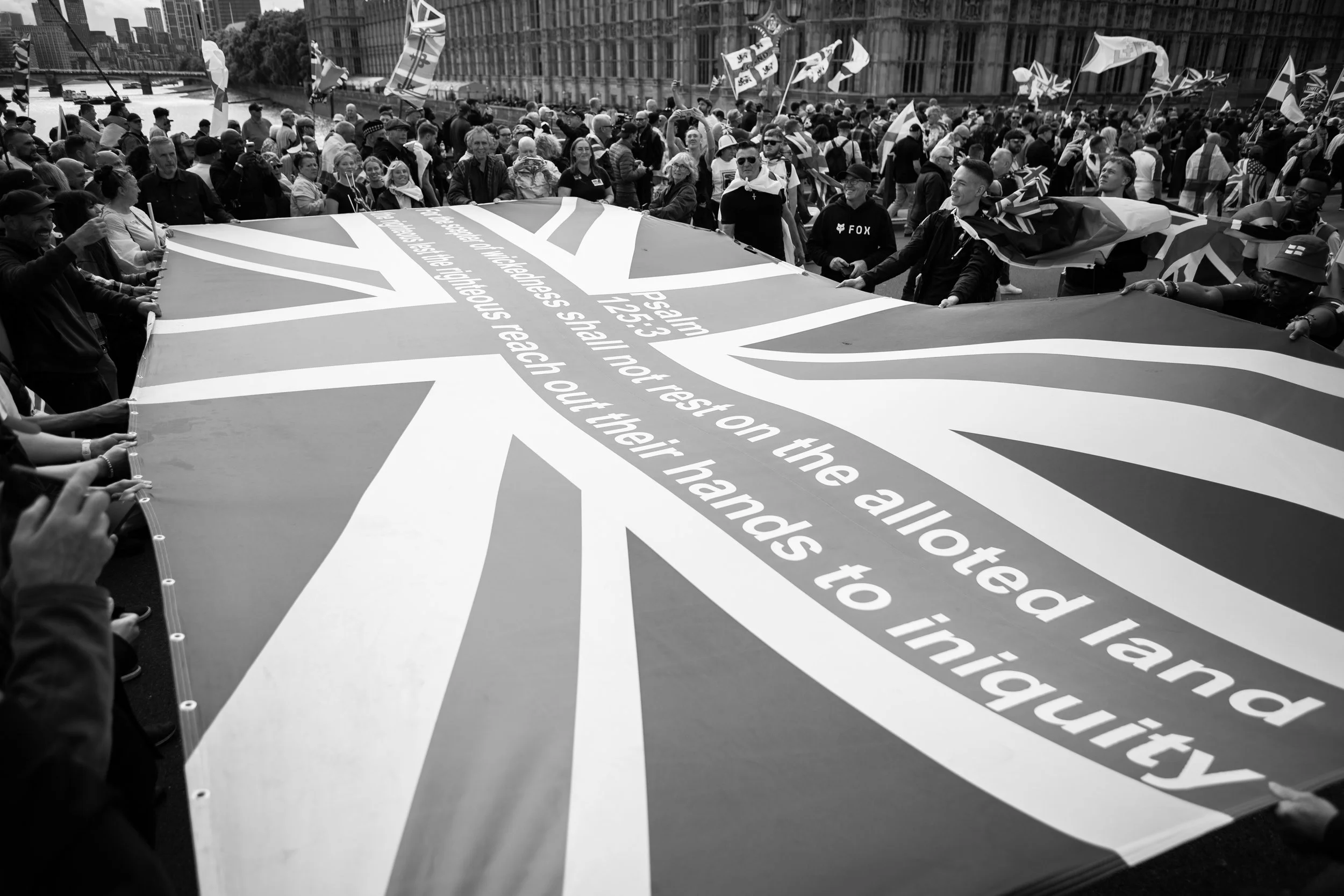 Crowd of people holding a large Union Jack flag with the lyrics from 'Imagine' by John Lennon on it, at a street protest or rally in an urban area.