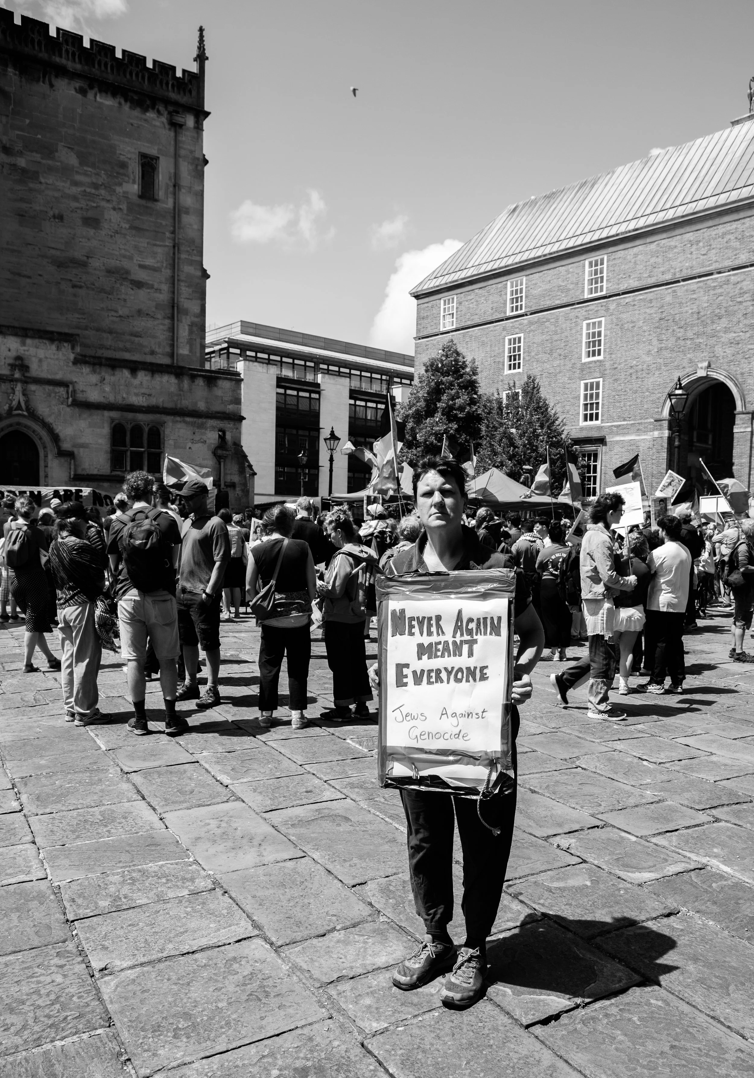 A woman holding a sign at a protest or rally with a crowd of people and flags in the background, in an urban setting with historical and modern buildings.