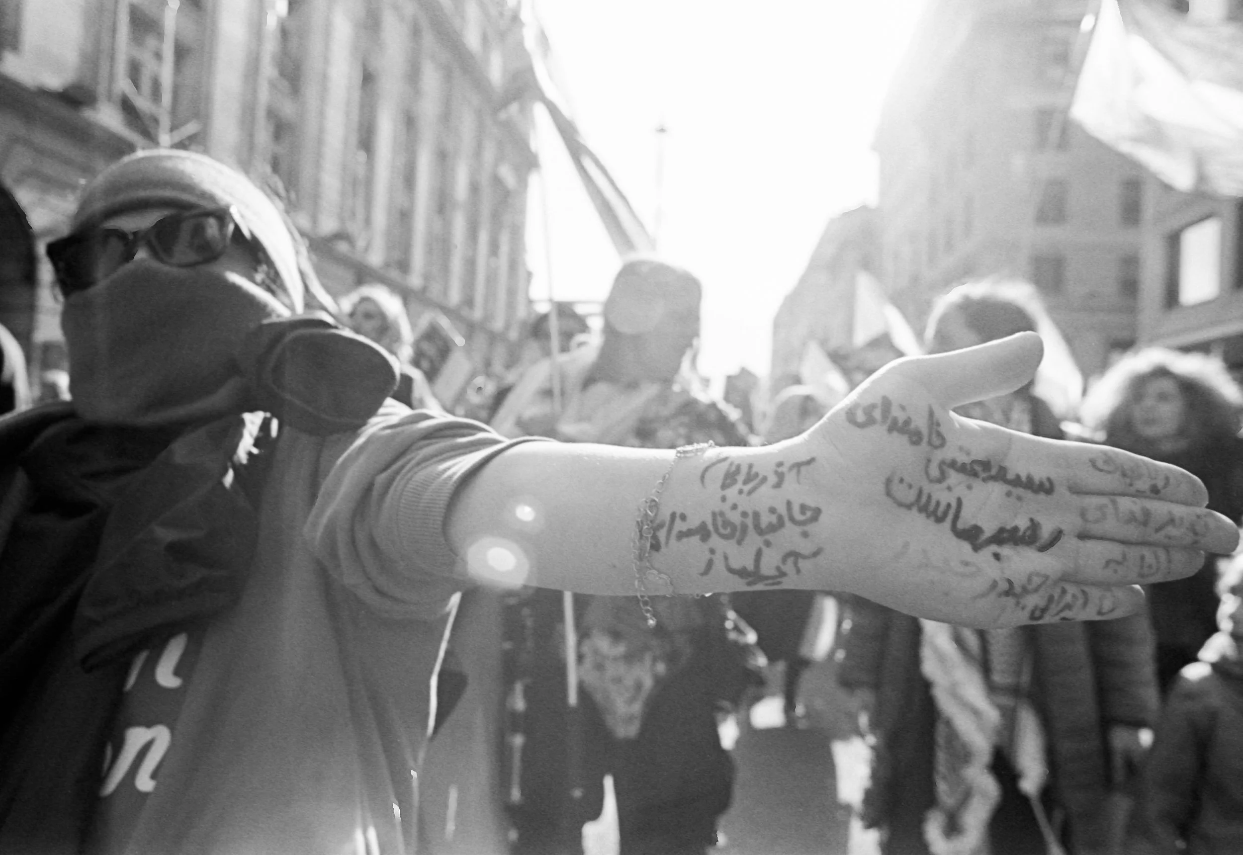 A person extending their arm forward with writing tattoos on their hand, in a crowded street scene with people and buildings, bright sunlight, and some flags or banners.