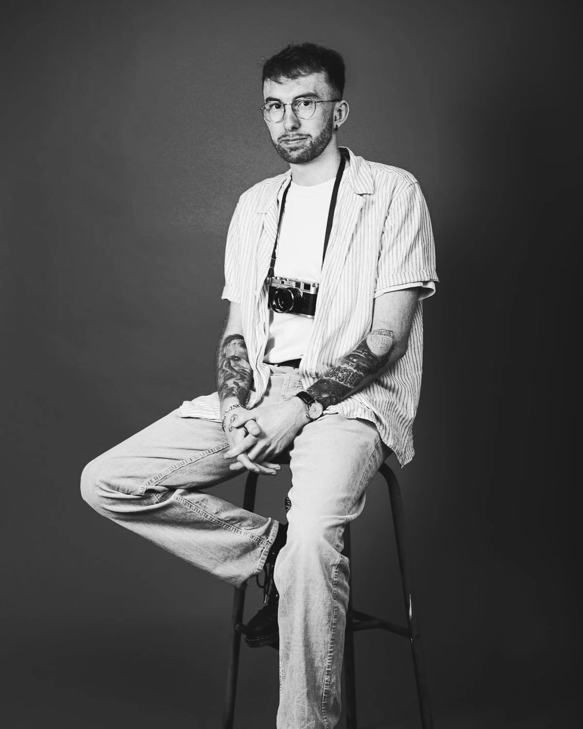 A young man with glasses and tattoos sitting on a stool, wearing a striped shirt, jeans, and a camera around his neck, in a black-and-white studio portrait.