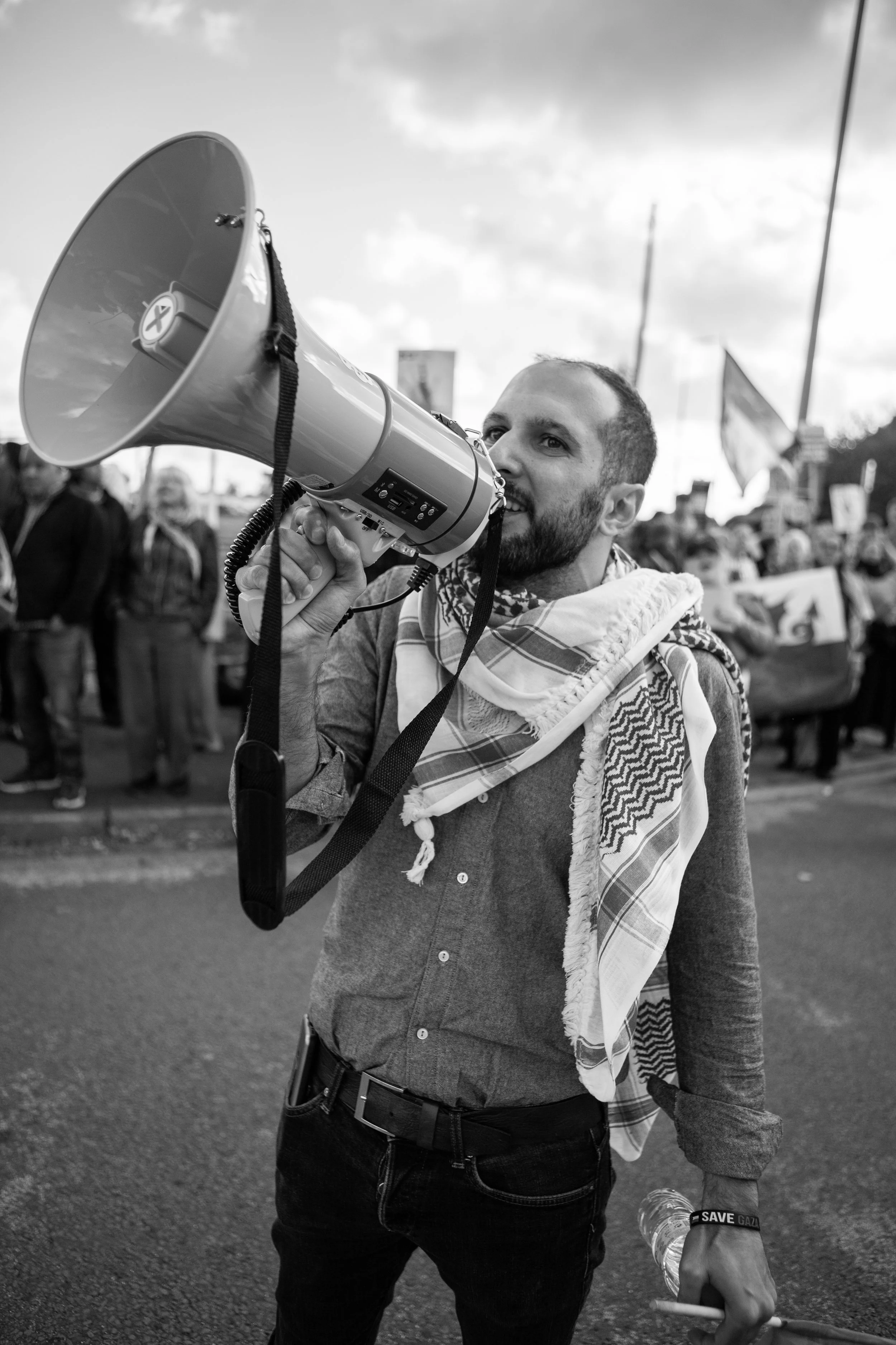 A man speaking into a megaphone at a protest or demonstration, with a crowd holding signs in the background.