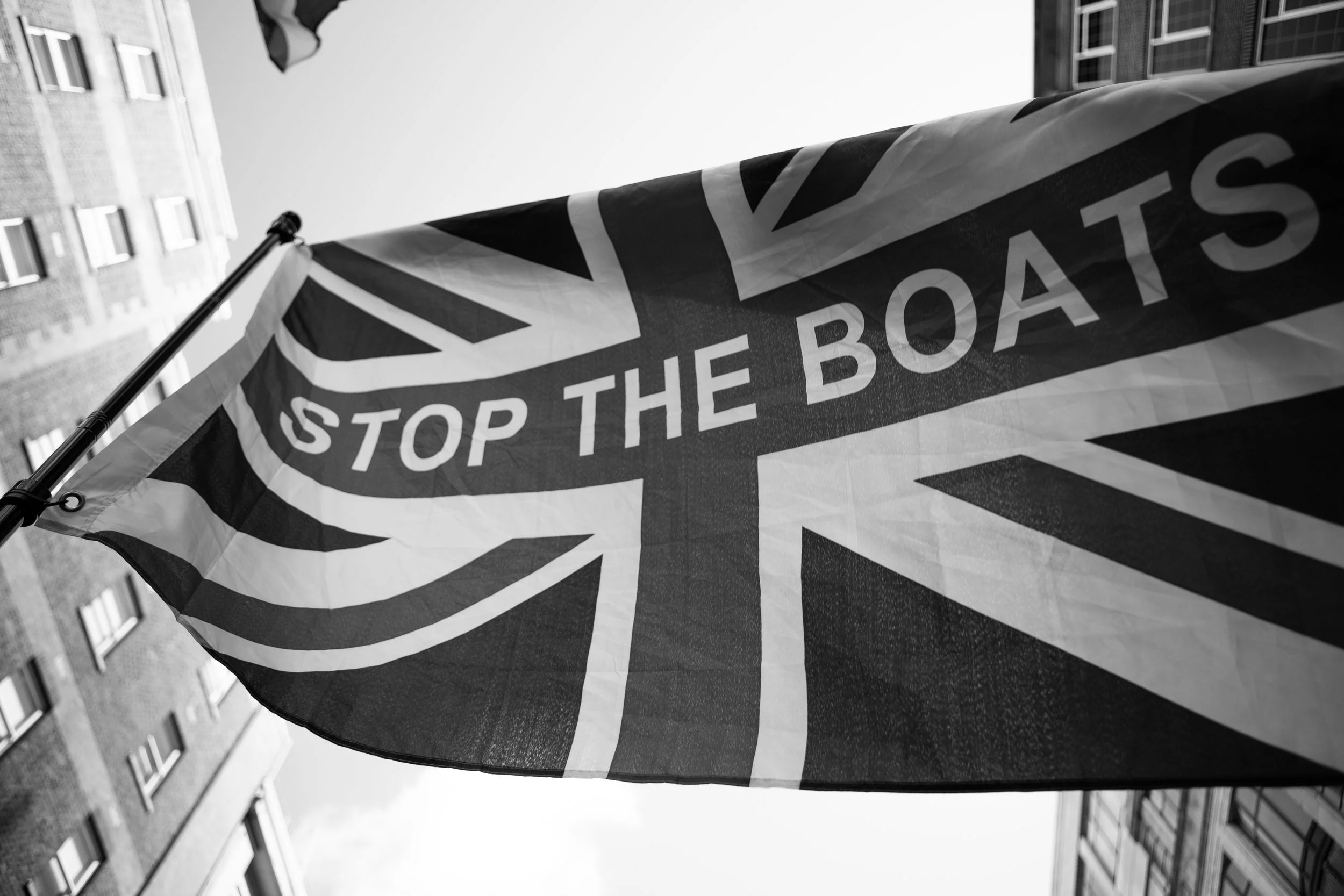 A flag with a Union Jack design and the words 'STOP THE BOATS' on it, flying outdoors with buildings in the background.