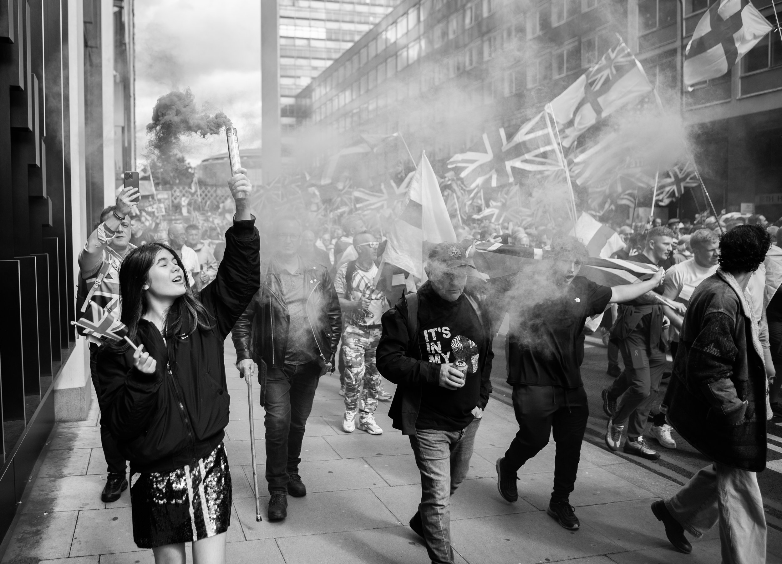 A black and white photo of a protest on a city street. A woman in the foreground holds a lit flare and a small Union Jack flag, with her eyes closed. Behind her, a large crowd of protesters carries multiple Union Jack flags. Smoke rises from the flar