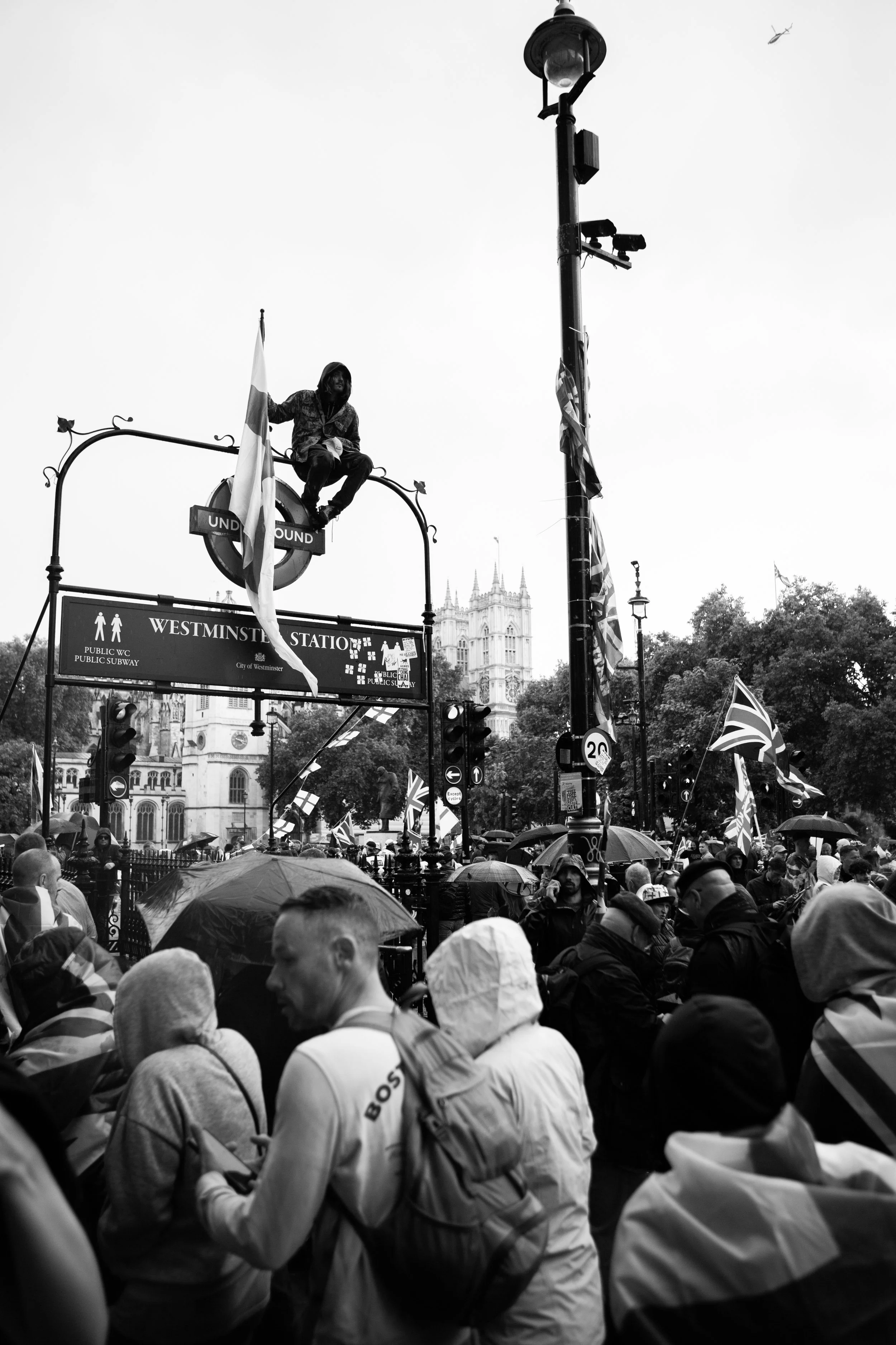 Crowd gathered outside Westminster Station with umbrellas, some people wearing hoods, many holding Union Jack flags. A figure with a hooded jacket sits on a sign hanging above the crowd, near a flagpole with a flag. The scene is overcast, with histor