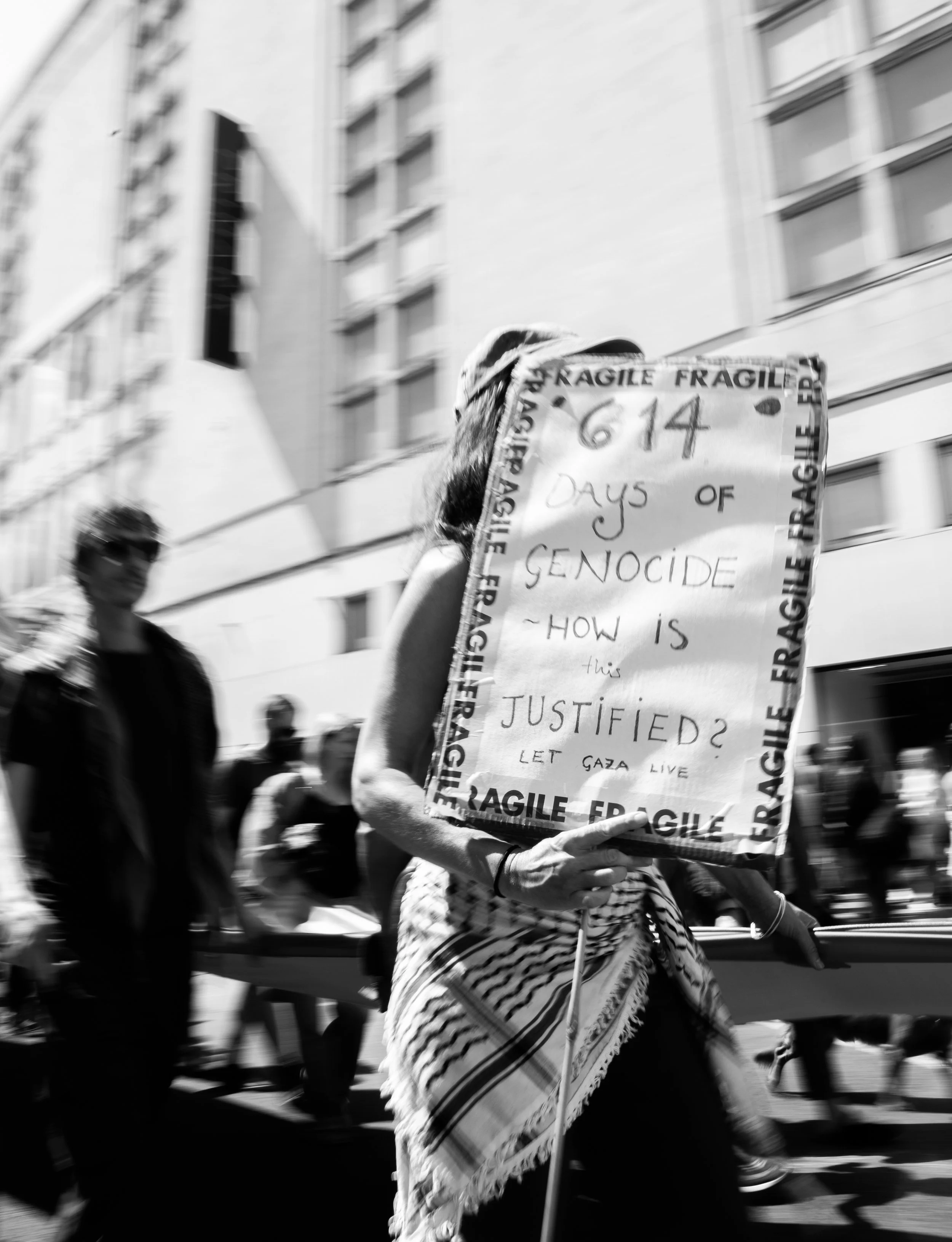 A person participating in a protest march holding a sign that reads '614 days of genocide - how is this justified? Let Gaza live.' The person wears a patterned scarf and a wristband, and other marchers are visible in the background on a city street.