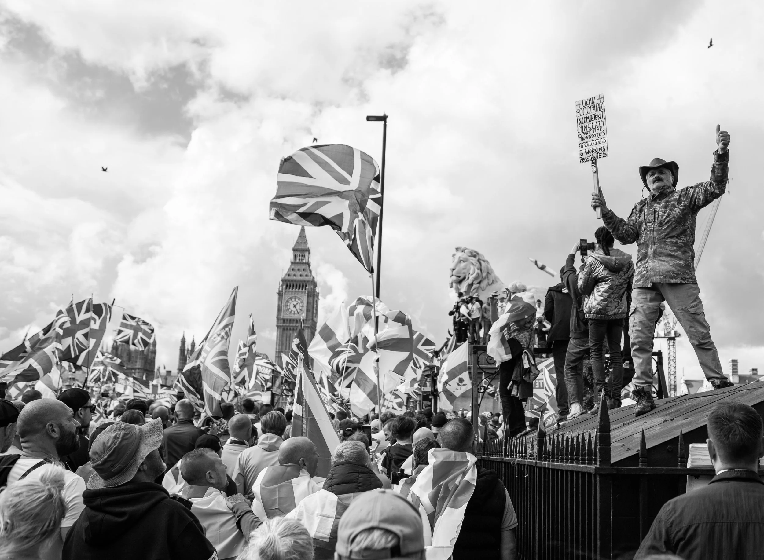 A large crowd of people gathered at a demonstration or protest near Big Ben, the clock tower in London. Many are holding Union Jack flags, and some are standing on a platform with a man holding a sign and giving a thumbs-up. The sky is partly cloudy 