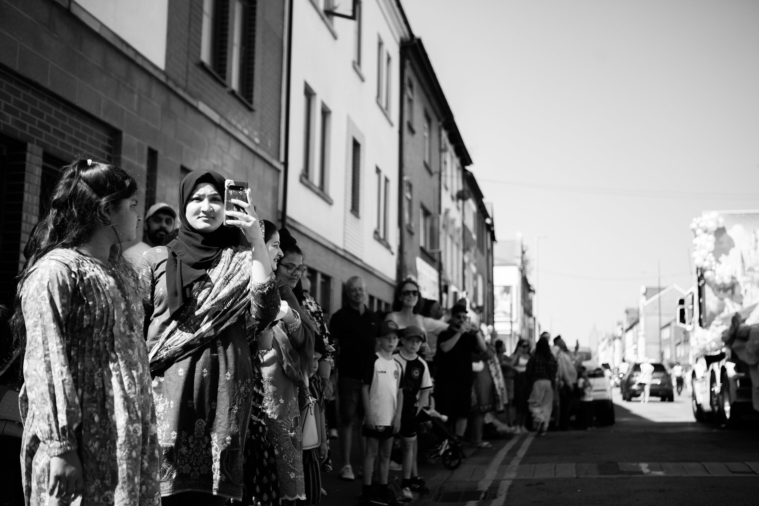 A crowd of people, including children and adults, lined up along a sidewalk on a city street, with some focused on a woman taking a photo or video with her phone. The scene is in black and white.