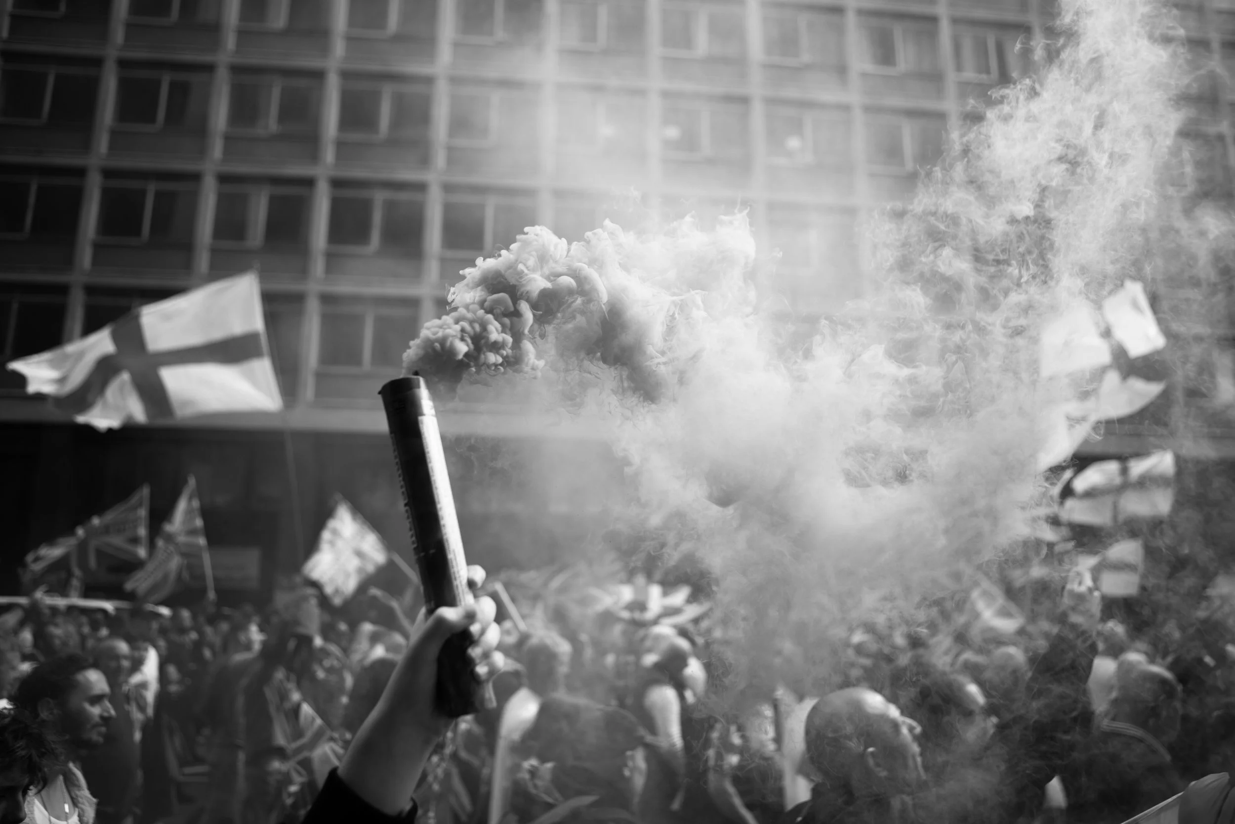 A black and white photo of a protest with a crowd of people, some holding flags, and a person in the foreground holding a flare emitting smoke.