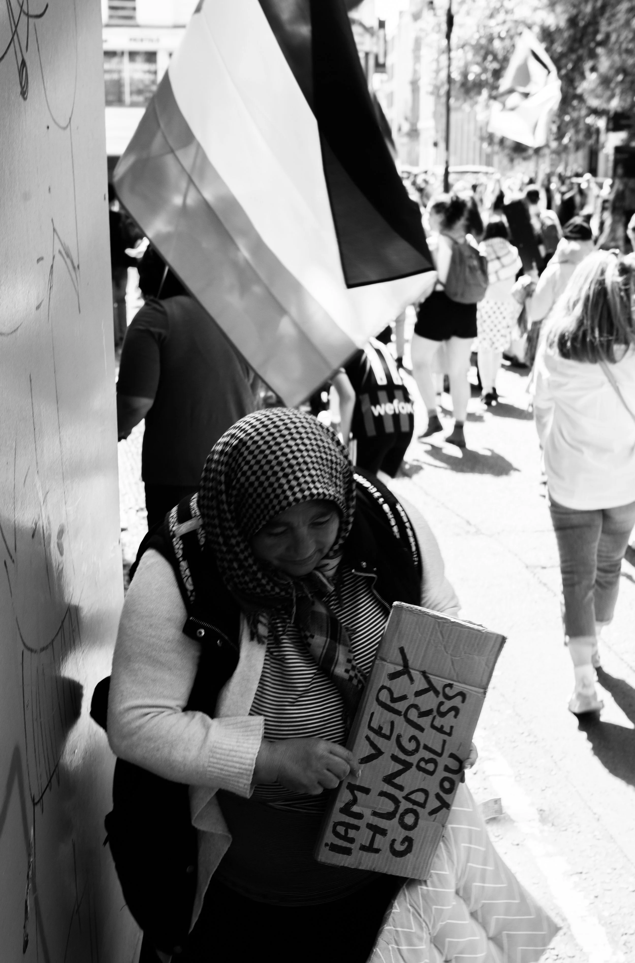 A woman in a headscarf holding a sign that says 'MANY ARE HUNGRY, GOD BLESS YOU' standing on a busy street with many pedestrians.