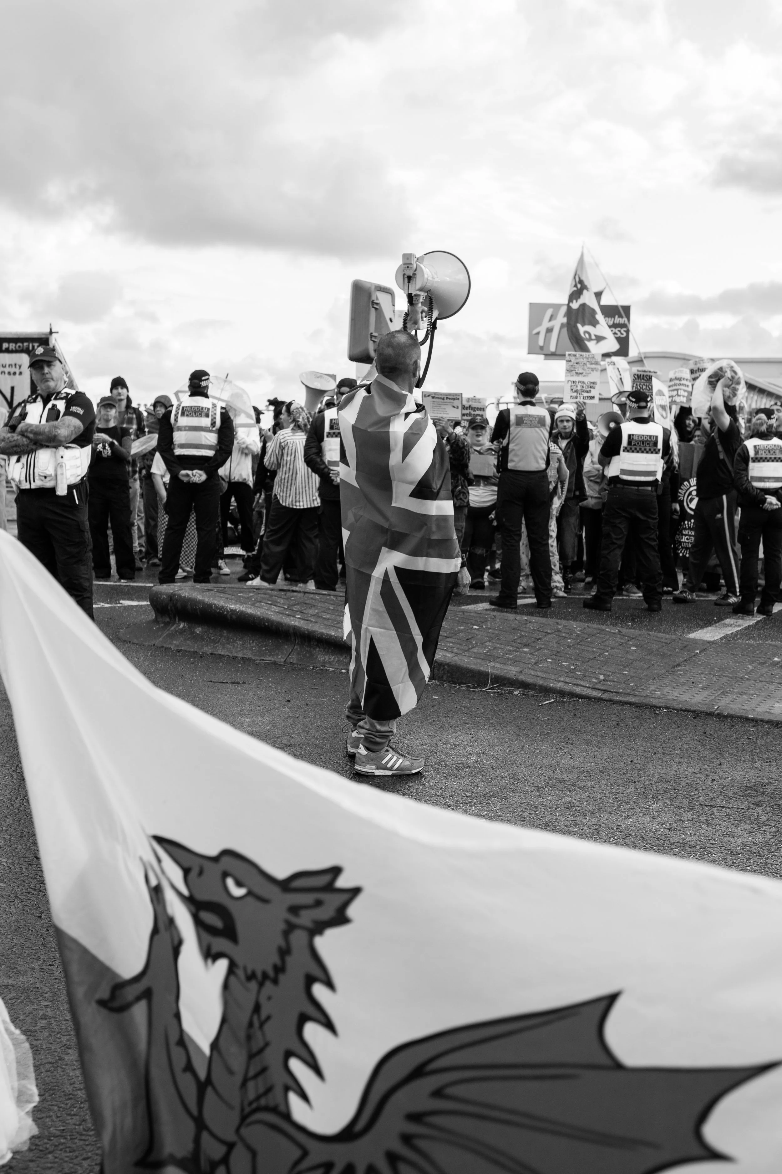 A man stands with his back to the camera, wrapped in a Welsh flag, facing a crowd of protesters and police officers at a demonstration, with protest signs and flags visible.