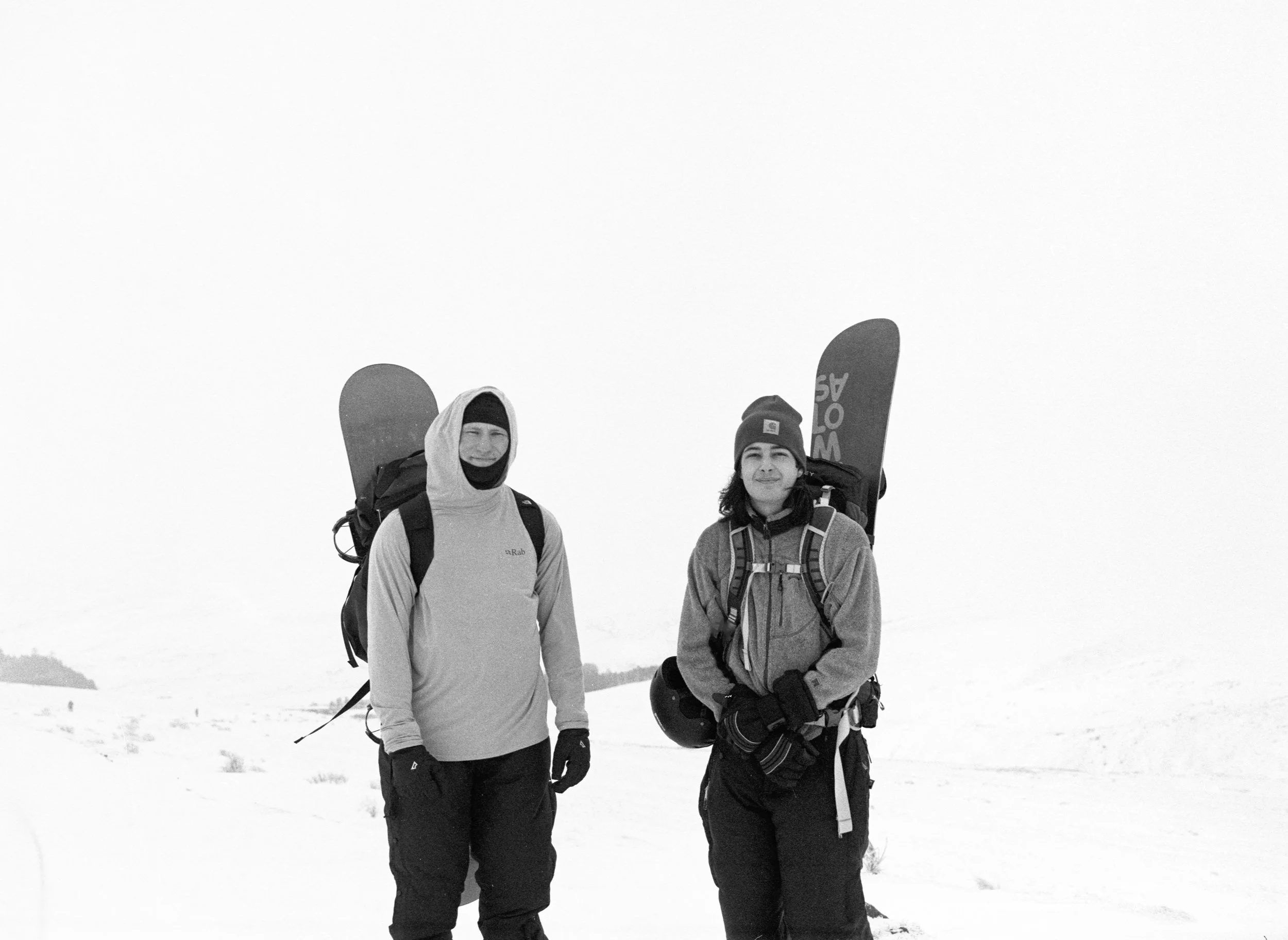 Two people standing in snowy landscape dressed in winter gear, each carrying snowboards on their backpacks.
