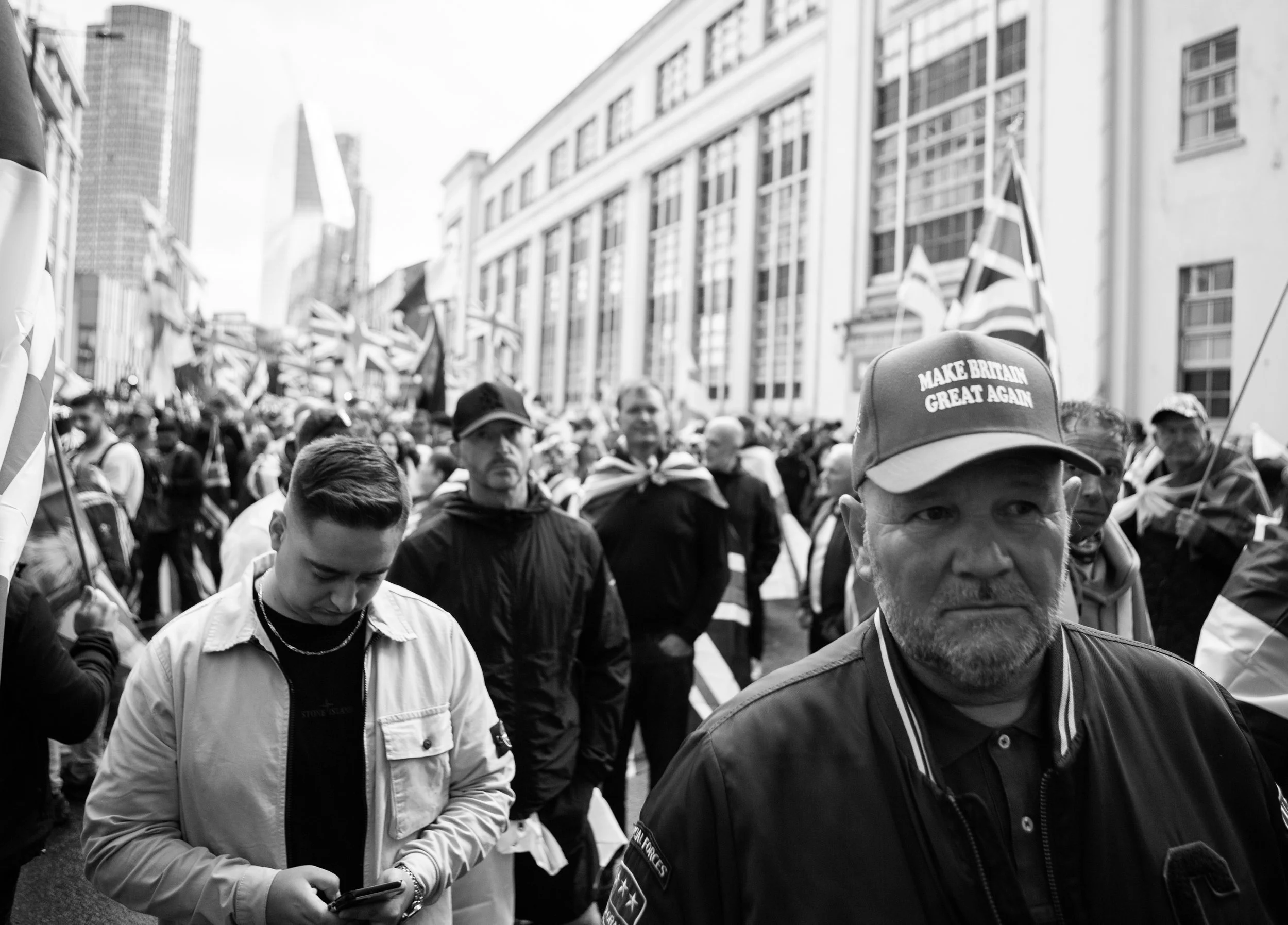 Crowd of people at a political demonstration holding British flags, one man in the foreground wearing a cap that reads 'Make Britain Great Again.'