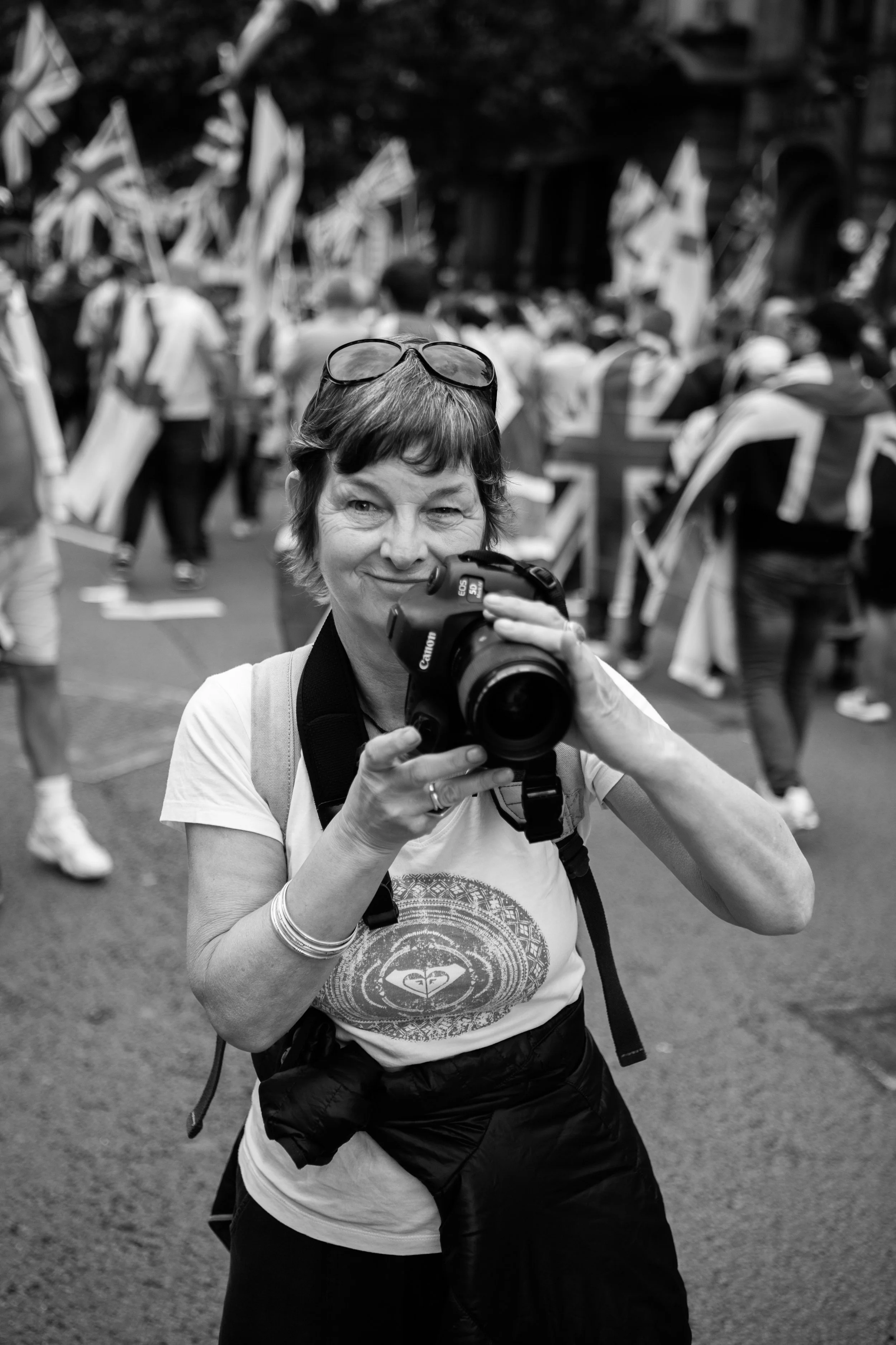 A woman holding a camera while taking a photo during a protest or demonstration, with people in the background carrying flags.