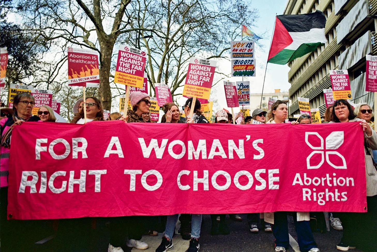 Women protesting with a large pink banner that reads 'For a woman's right to choose' and 'Abortion Rights'. Many women hold signs advocating for women's rights, abortion access, and anti-racism, with some signs saying 'Abortion is healthcare' and 'Wo