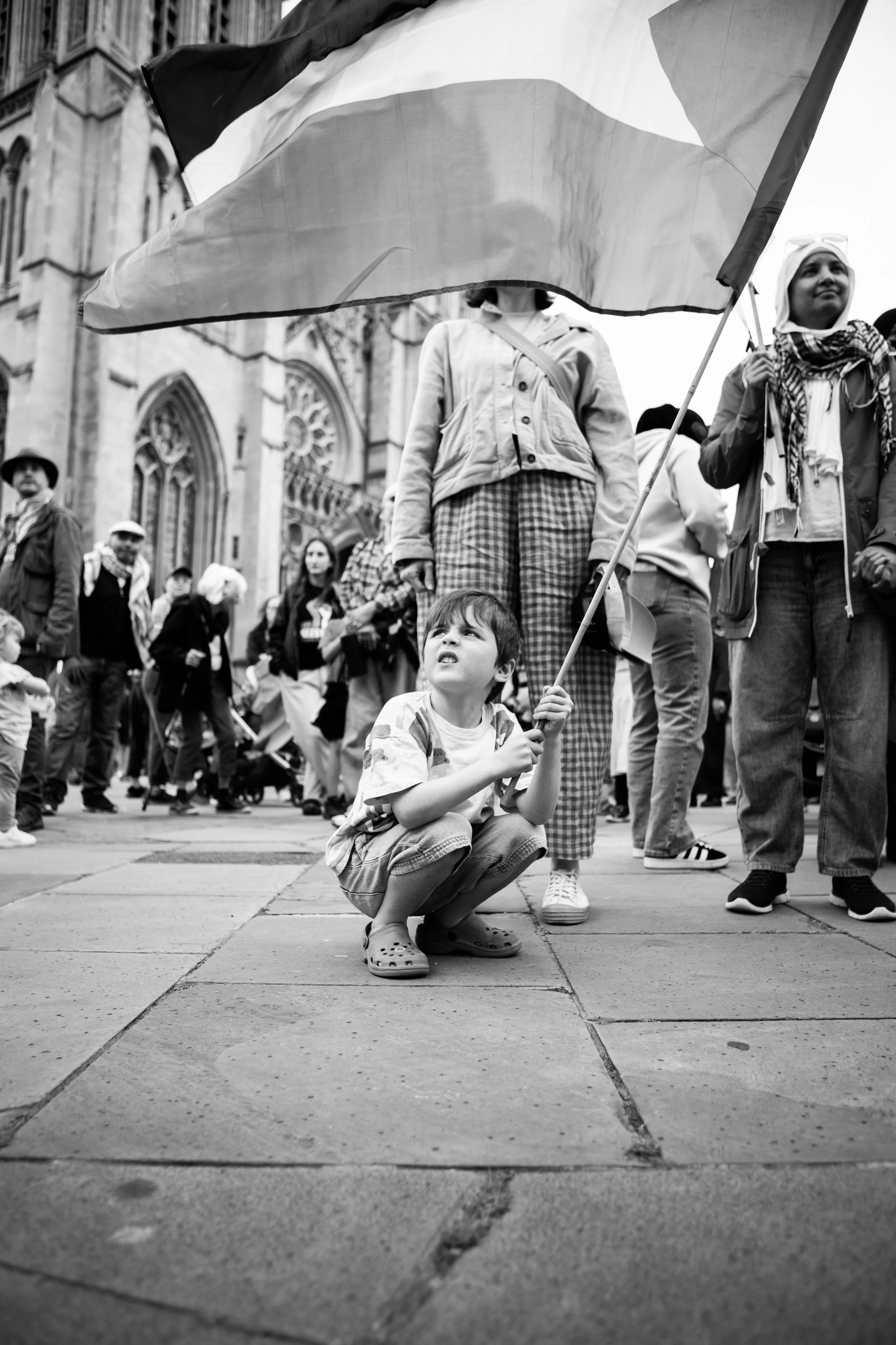 A young boy squatting on the sidewalk, holding a flag, during a crowded outdoor event in front of a historic building.
