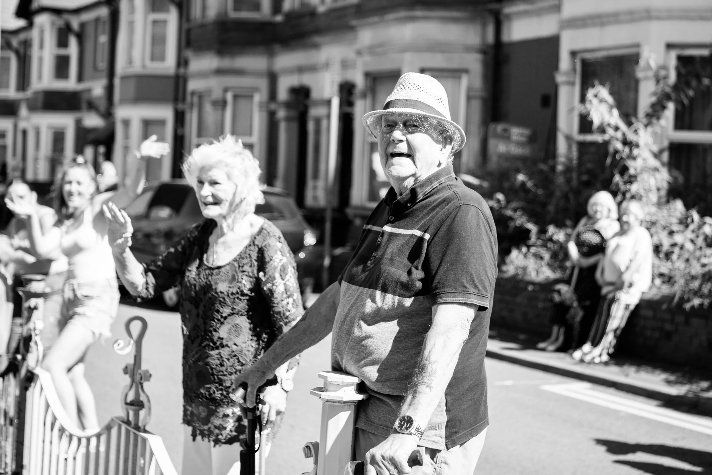 Older man wearing a hat and sunglasses walking outdoors beside an elderly woman, with several people in the background on a sunny day.