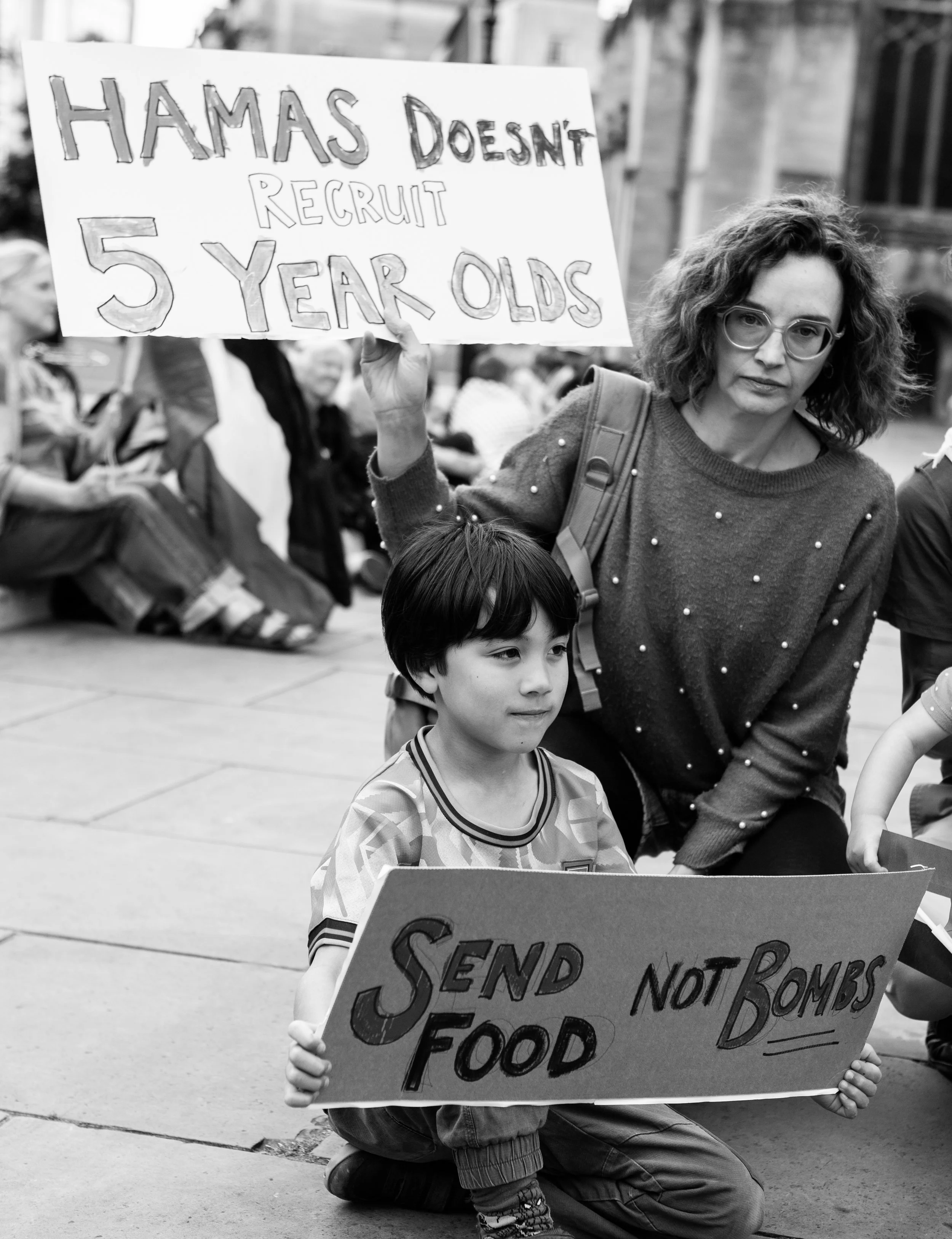 A woman and a boy holding protest signs. The woman’s sign says, 'Hamas doesn’t recruit 5 year olds.' The boy’s sign says, 'Send food not bombs.' They are participating in a demonstration or protest, with other people sitting and standing in the backg